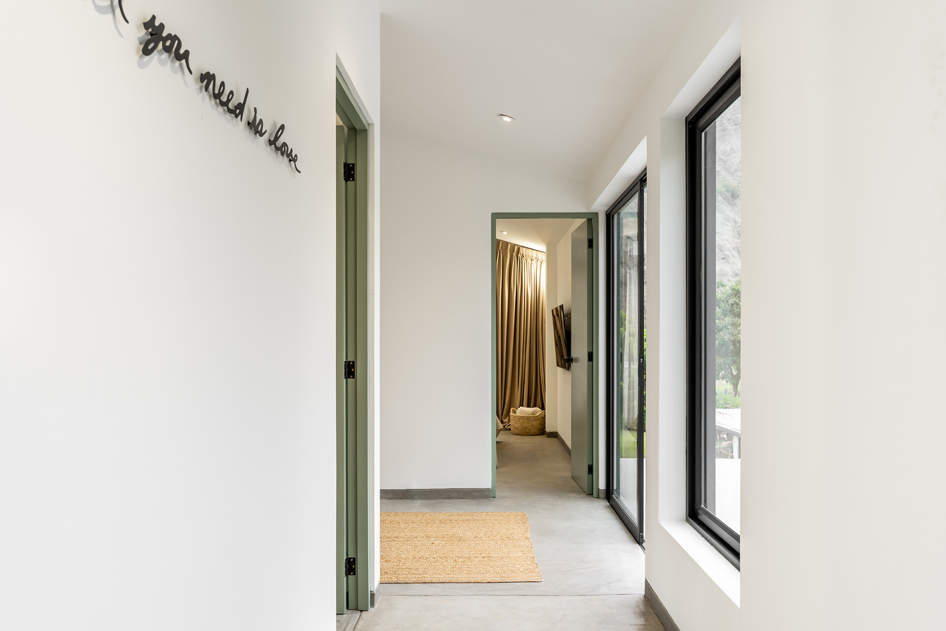 Casa Berna’s upstairs hallway pairs white walls and a concrete floor with pale green doorframes and tall windows overlooking the greenery outside.
