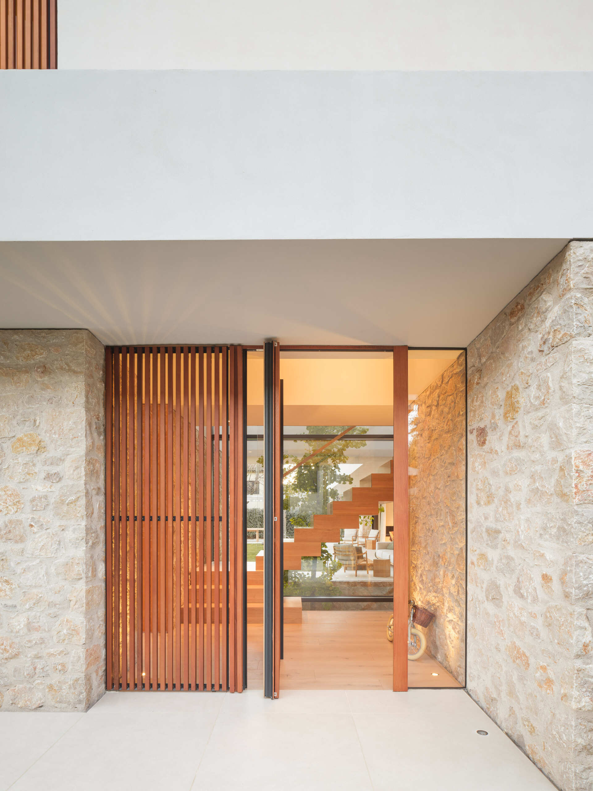 A sheltered glass front door opens into a bright entrance with pale timber flooring, exposed stone, and a wooden staircase. Discover the entrance design at House 2194 by Nordest Architecture SLP in Olot, Spain.