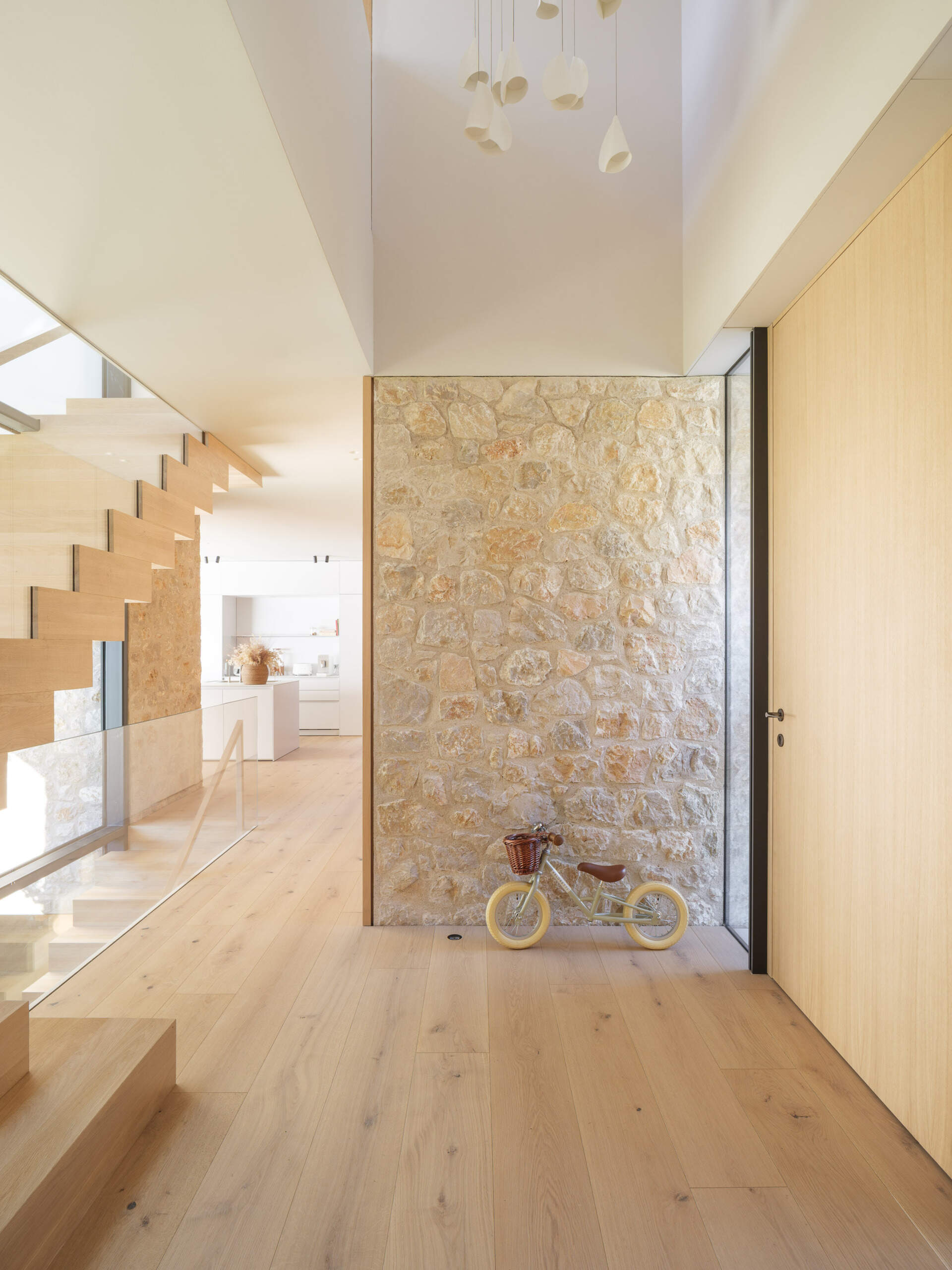 A sheltered glass front door opens into a bright entrance with pale timber flooring, exposed stone, and a wooden staircase. Discover the entrance design at House 2194 by Nordest Architecture SLP in Olot, Spain.