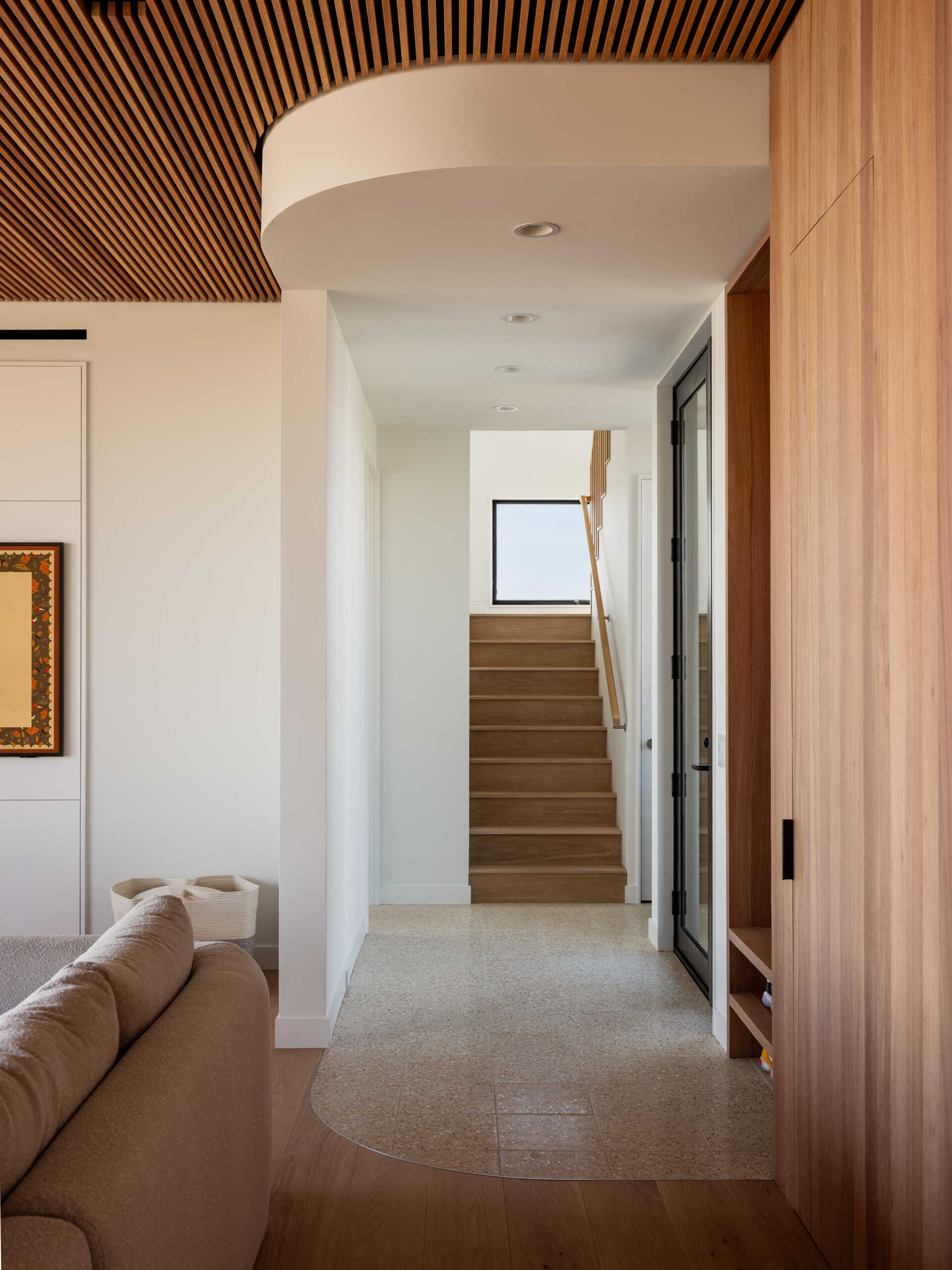 Wood storage, black coat hooks, and open shoe shelves make this Fire Island entryway practical and welcoming. A curved white soffit and rounded tile border add softness beneath a slatted wood ceiling.