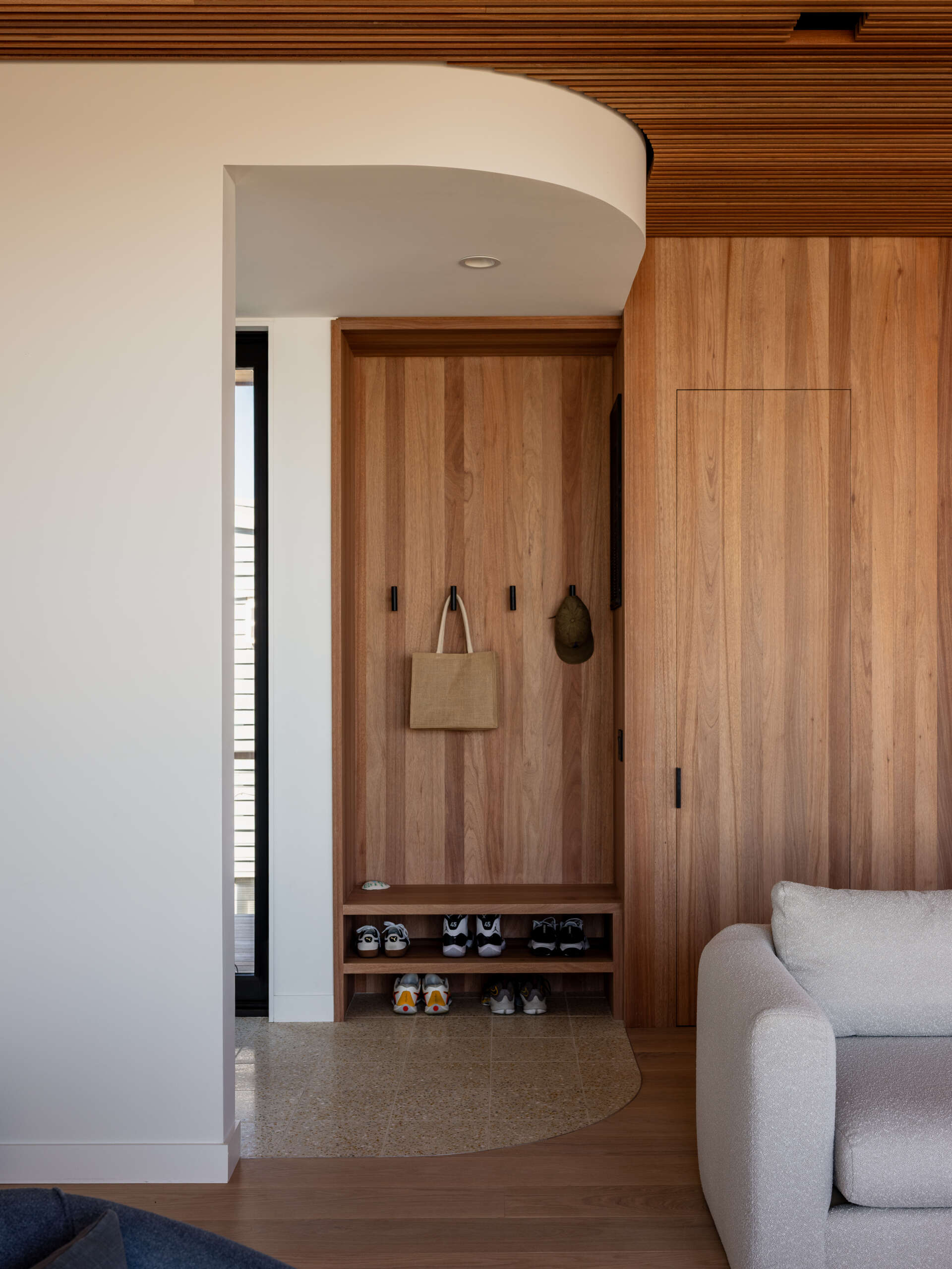 Wood storage, black coat hooks, and open shoe shelves make this Fire Island entryway practical and welcoming. A curved white soffit and rounded tile border add softness beneath a slatted wood ceiling.