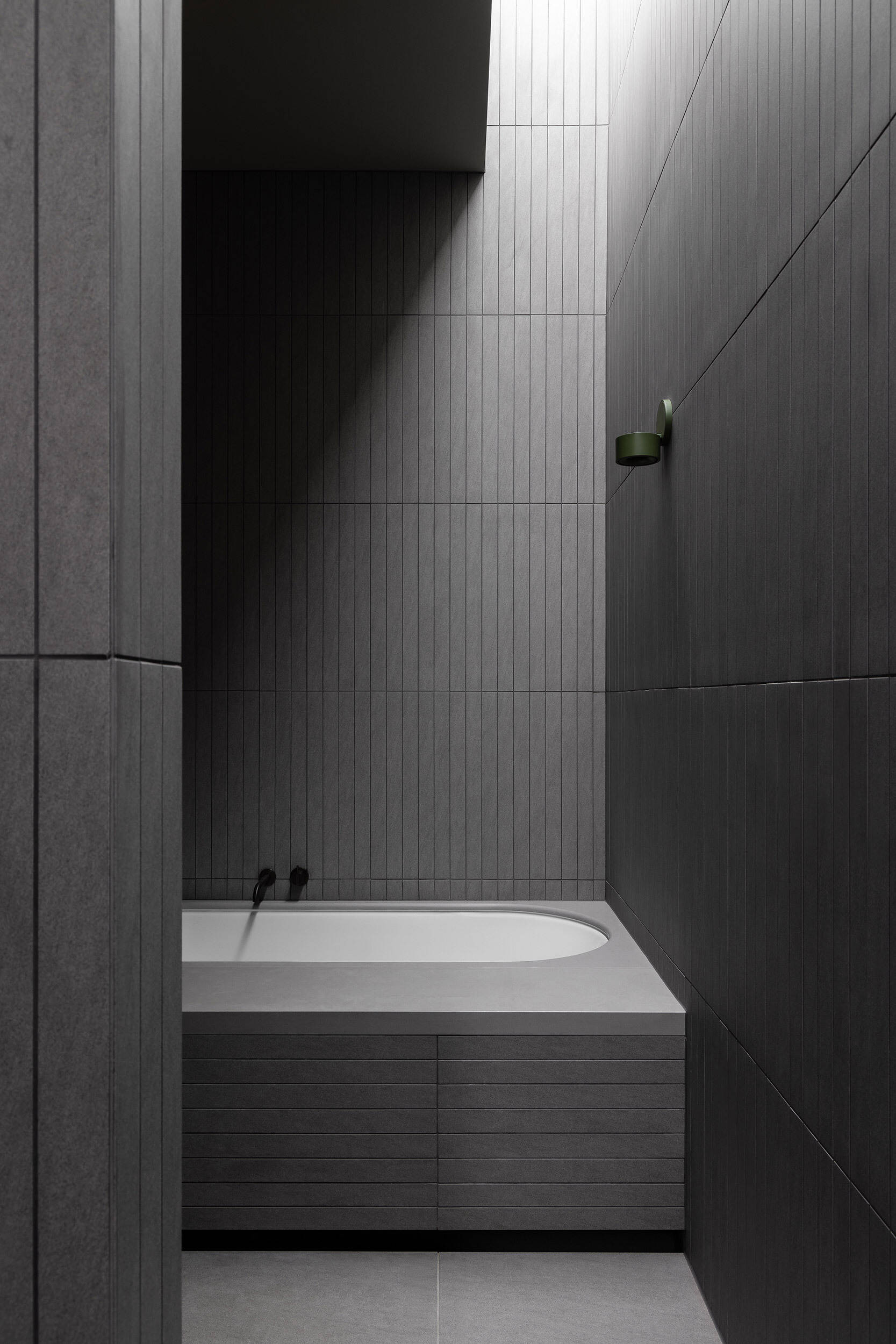 This modern bathroom is lined in slender charcoal tiles around a built-in bath. Light enters from above, catching the vertical tile joints and giving the room a more enclosed, dramatic feel while still keeping the material palette restrained.