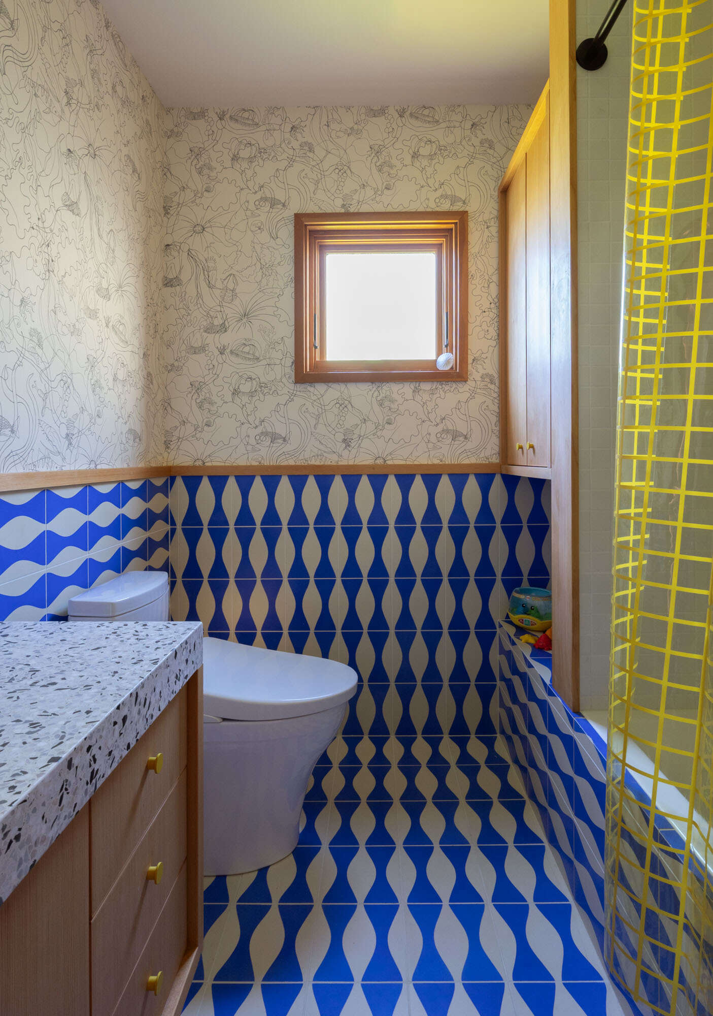 See the guest bathroom before and after, transformed from a neutral space into a playful blue room with wave-pattern tile, sea creature wallpaper, pale timber cabinetry, terrazzo and a translucent yellow shower curtain.