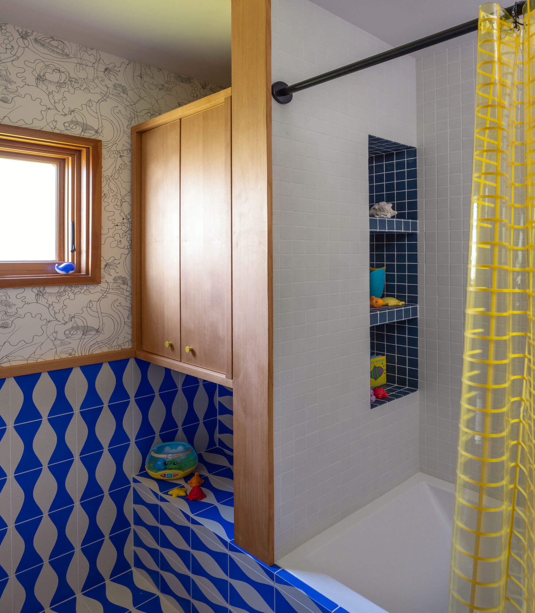 See the guest bathroom before and after, transformed from a neutral space into a playful blue room with wave-pattern tile, sea creature wallpaper, pale timber cabinetry, terrazzo and a translucent yellow shower curtain.