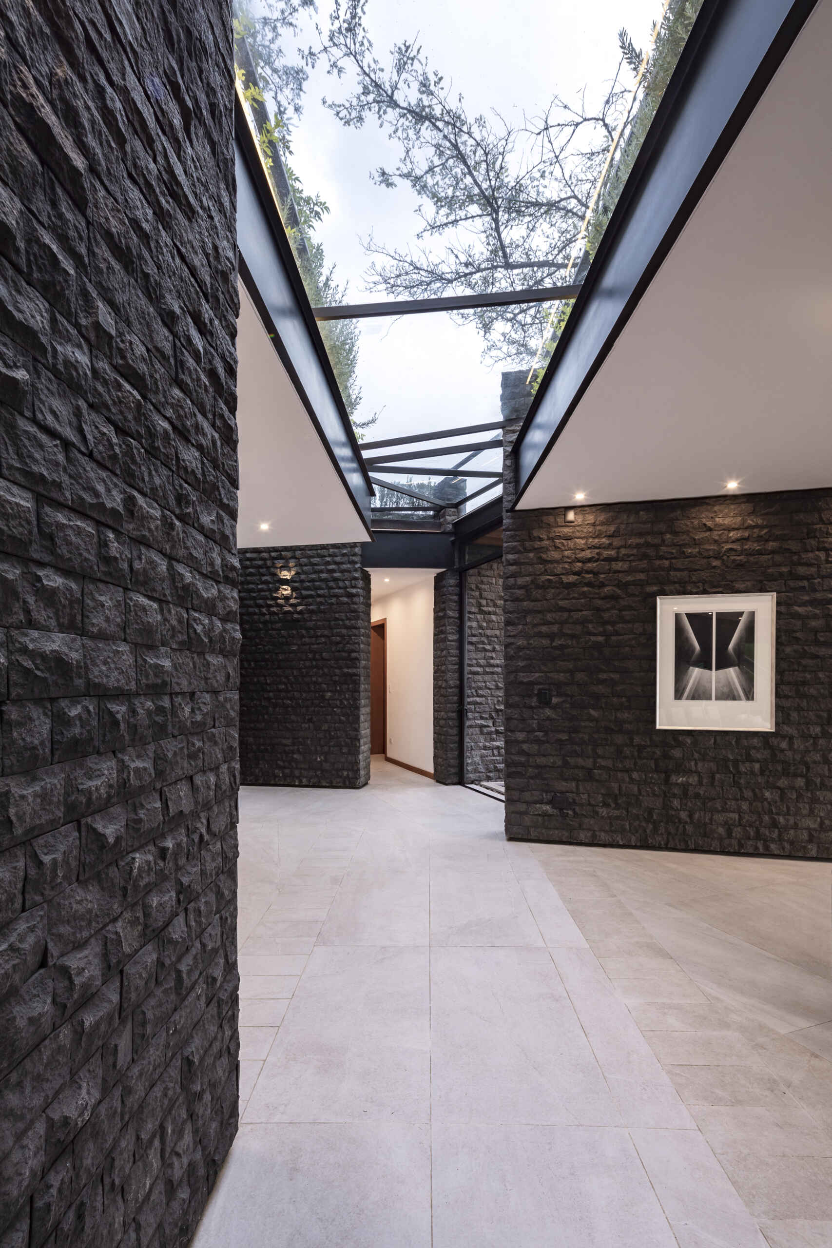 Explore the Hidden Stone House hallway, where dark stone walls, black steel framing, skylights and views of the trees create a dramatic architectural passage.