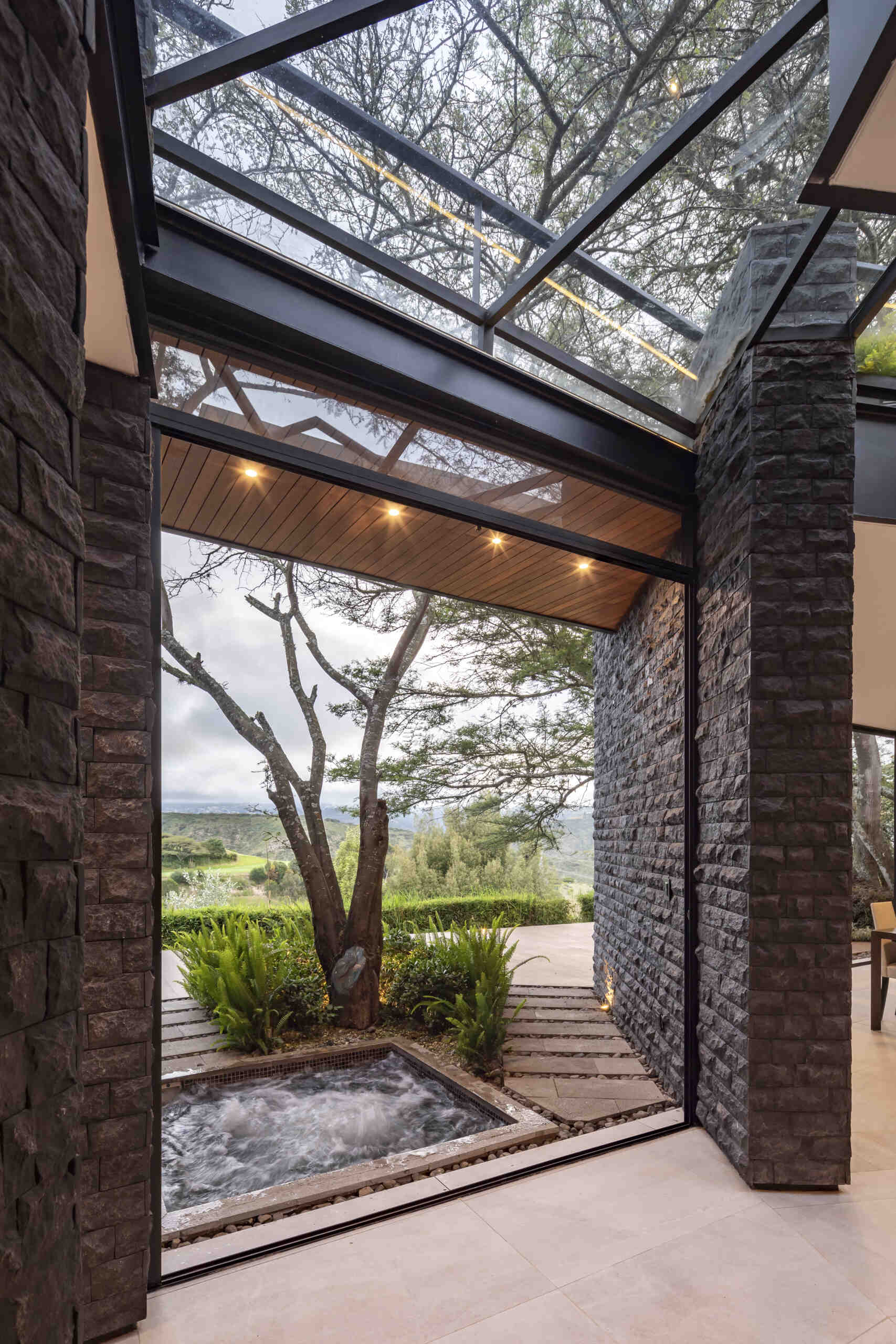 Explore the Hidden Stone House hallway, where dark stone walls, black steel framing, skylights and views of the trees create a dramatic architectural passage.