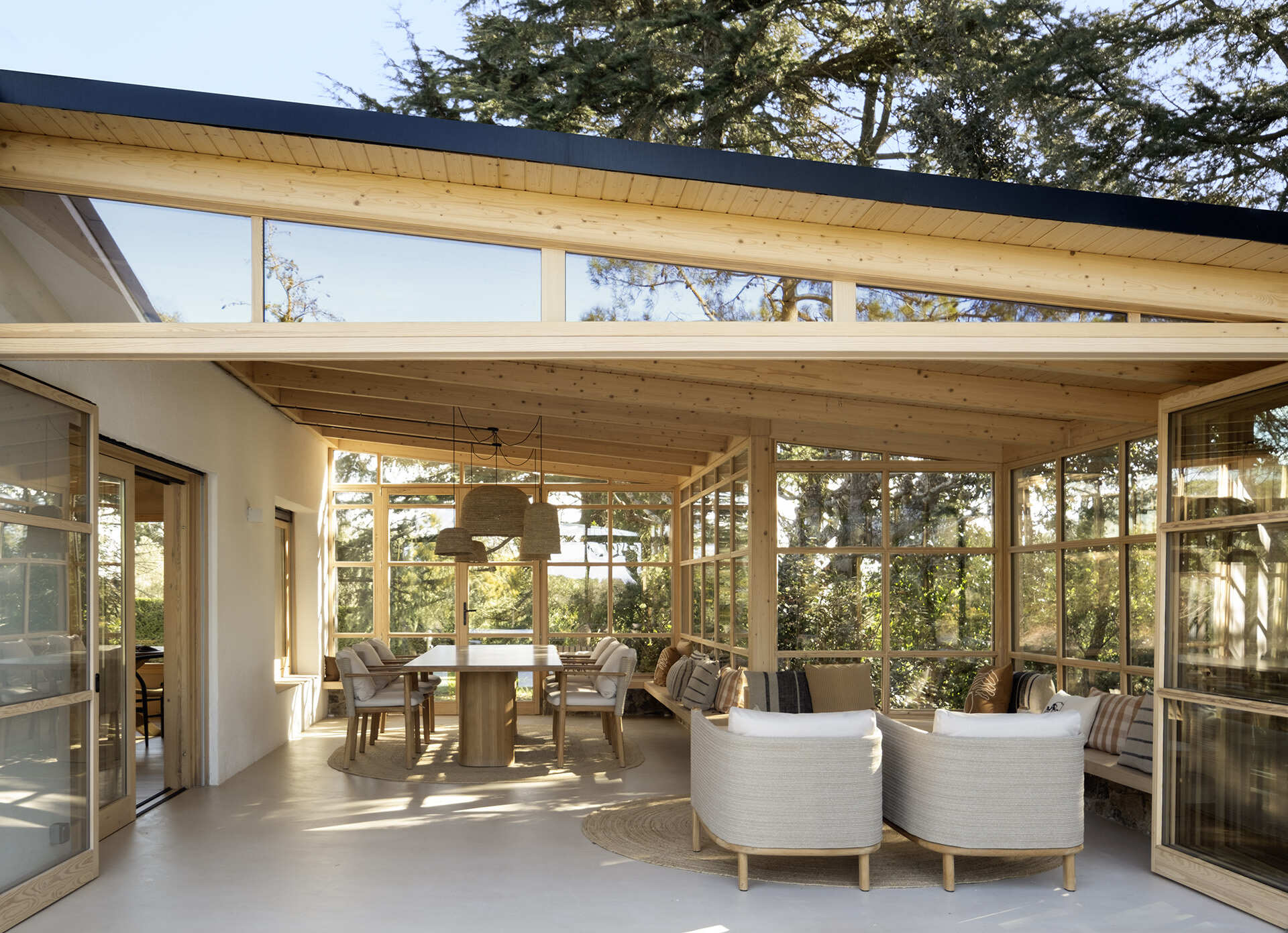 This glass-wrapped greenhouse is one of the most inviting spaces in the home, combining a long dining table, woven pendant lights and a relaxed lounge surrounded by trees. Timber framing and built-in seating make the forest feel almost within reach.