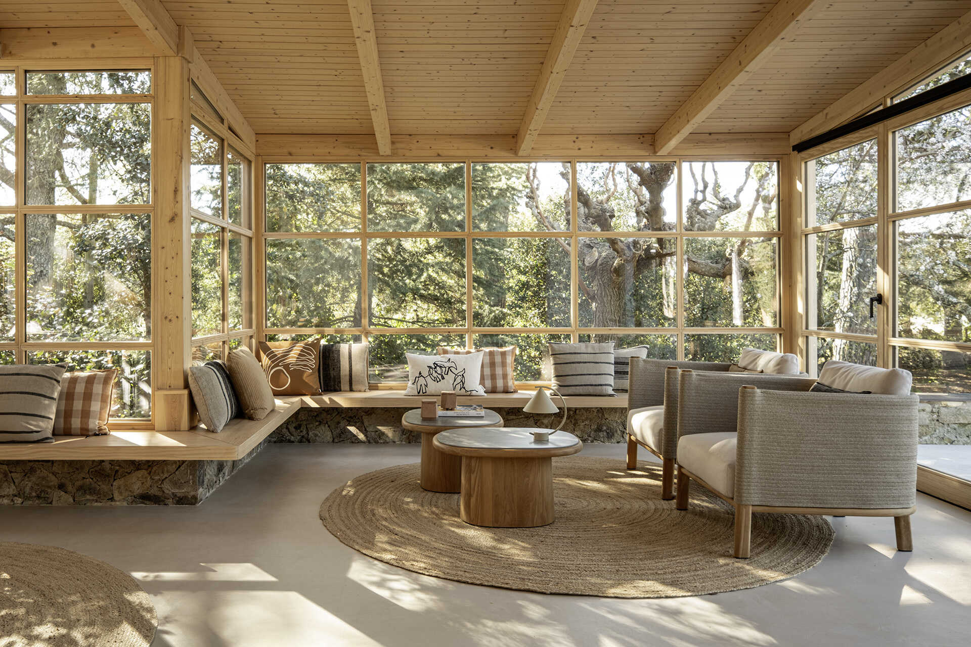 This glass-wrapped greenhouse is one of the most inviting spaces in the home, combining a long dining table, woven pendant lights and a relaxed lounge surrounded by trees. Timber framing and built-in seating make the forest feel almost within reach.