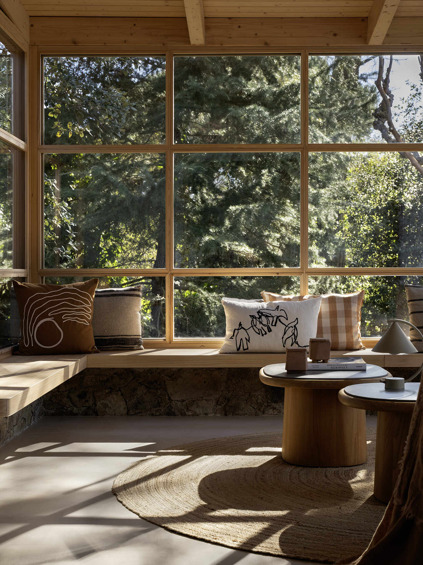 This glass-wrapped greenhouse is one of the most inviting spaces in the home, combining a long dining table, woven pendant lights and a relaxed lounge surrounded by trees. Timber framing and built-in seating make the forest feel almost within reach.