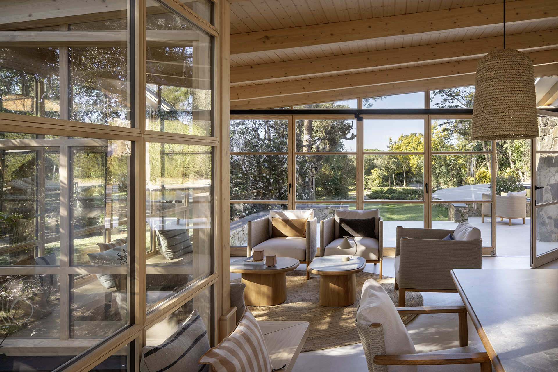 This glass-wrapped greenhouse is one of the most inviting spaces in the home, combining a long dining table, woven pendant lights and a relaxed lounge surrounded by trees. Timber framing and built-in seating make the forest feel almost within reach.