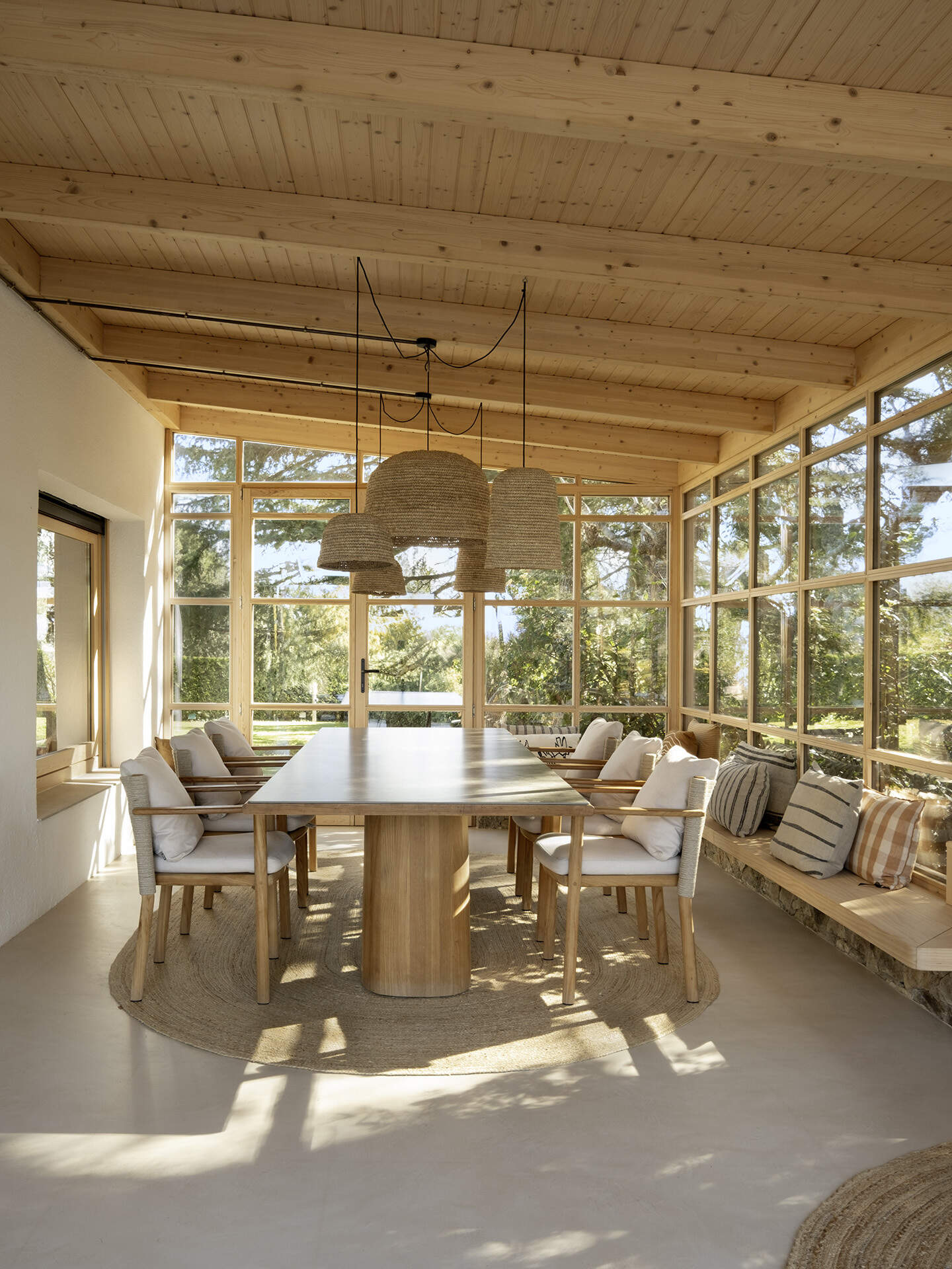 This glass-wrapped greenhouse is one of the most inviting spaces in the home, combining a long dining table, woven pendant lights and a relaxed lounge surrounded by trees. Timber framing and built-in seating make the forest feel almost within reach.