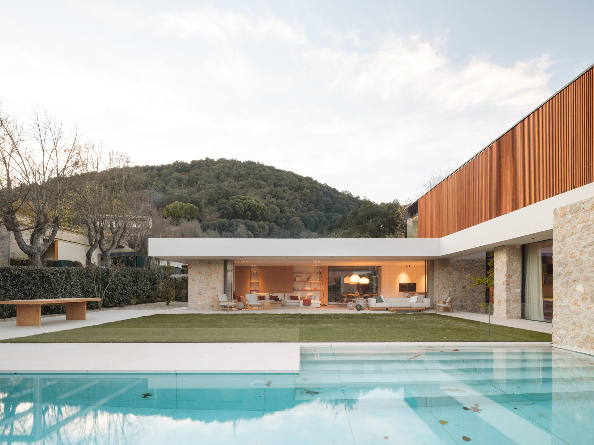 Stone walls, wooden screens, and a projecting concrete slab define this contemporary home in Olot, Spain. Explore House 2194 by Nordest Architecture SLP, with an L-shaped layout, a sheltered garden, and a layered façade.