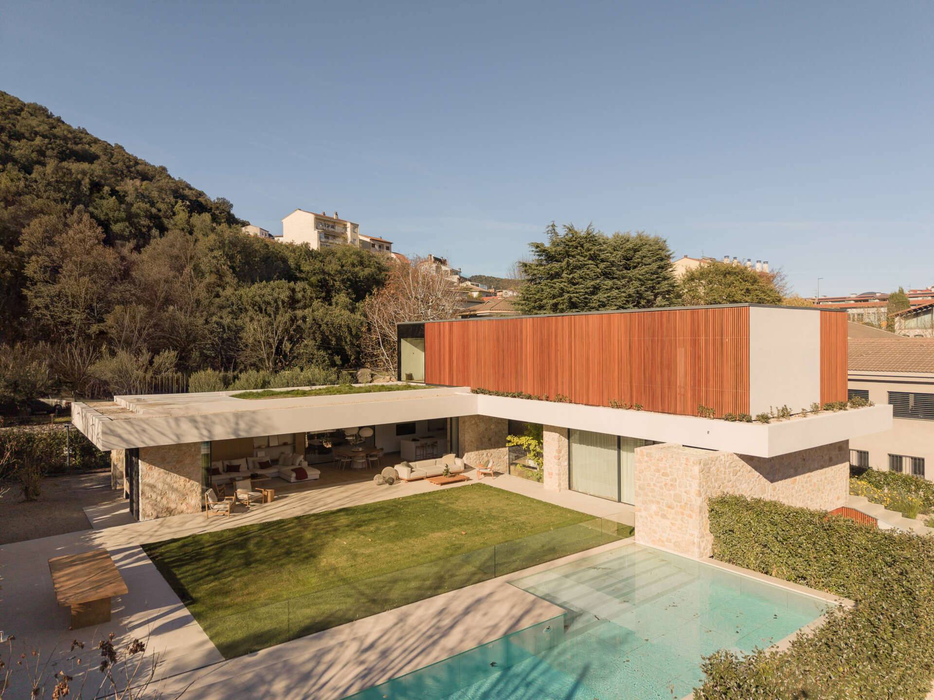 A shaded patio, timber outdoor furniture, central lawn, and pool create an inviting backyard at House 2194 in Olot, Spain. Sliding glass doors open the living and dining spaces directly onto the garden.