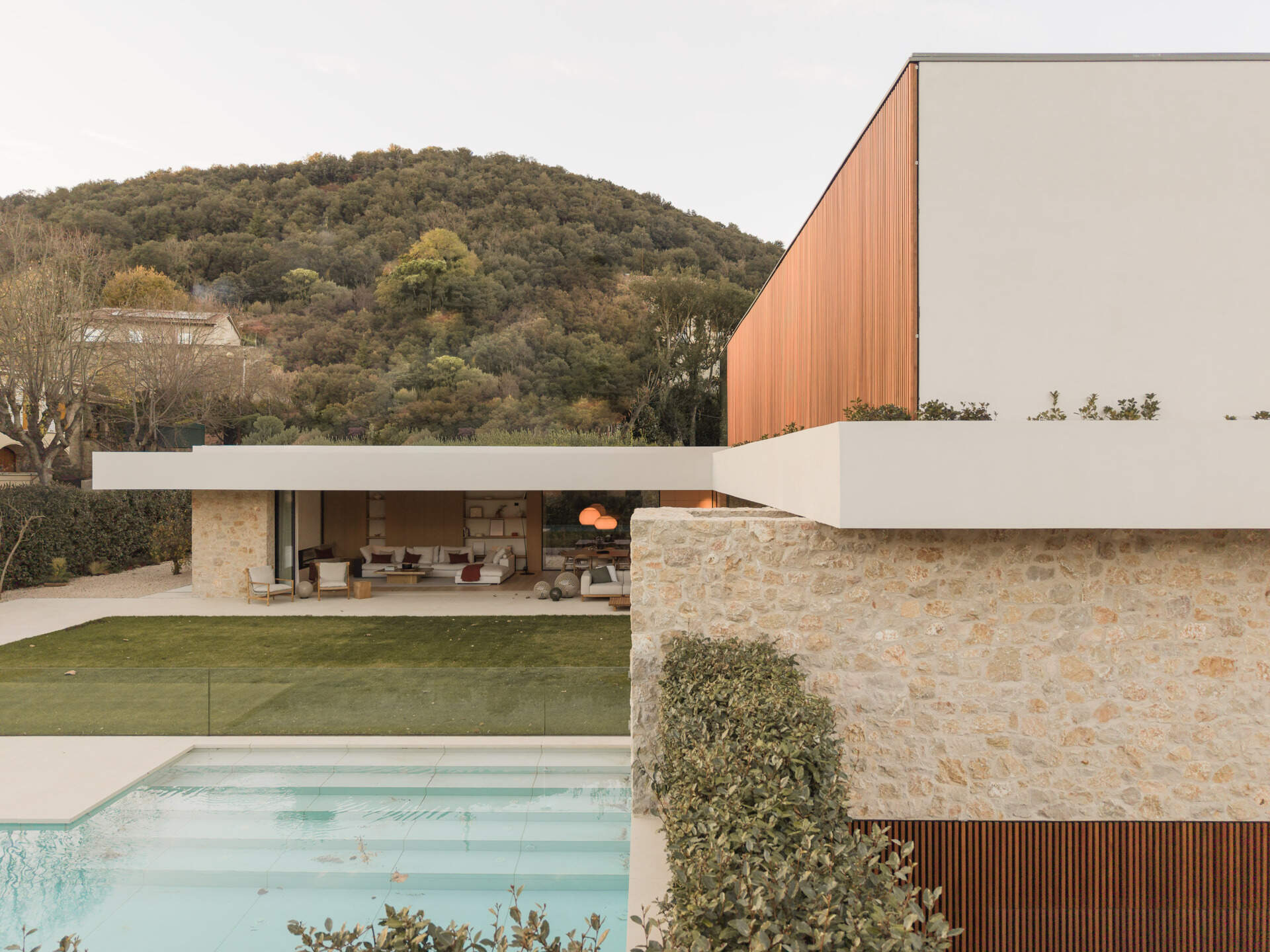 A shaded patio, timber outdoor furniture, central lawn, and pool create an inviting backyard at House 2194 in Olot, Spain. Sliding glass doors open the living and dining spaces directly onto the garden.