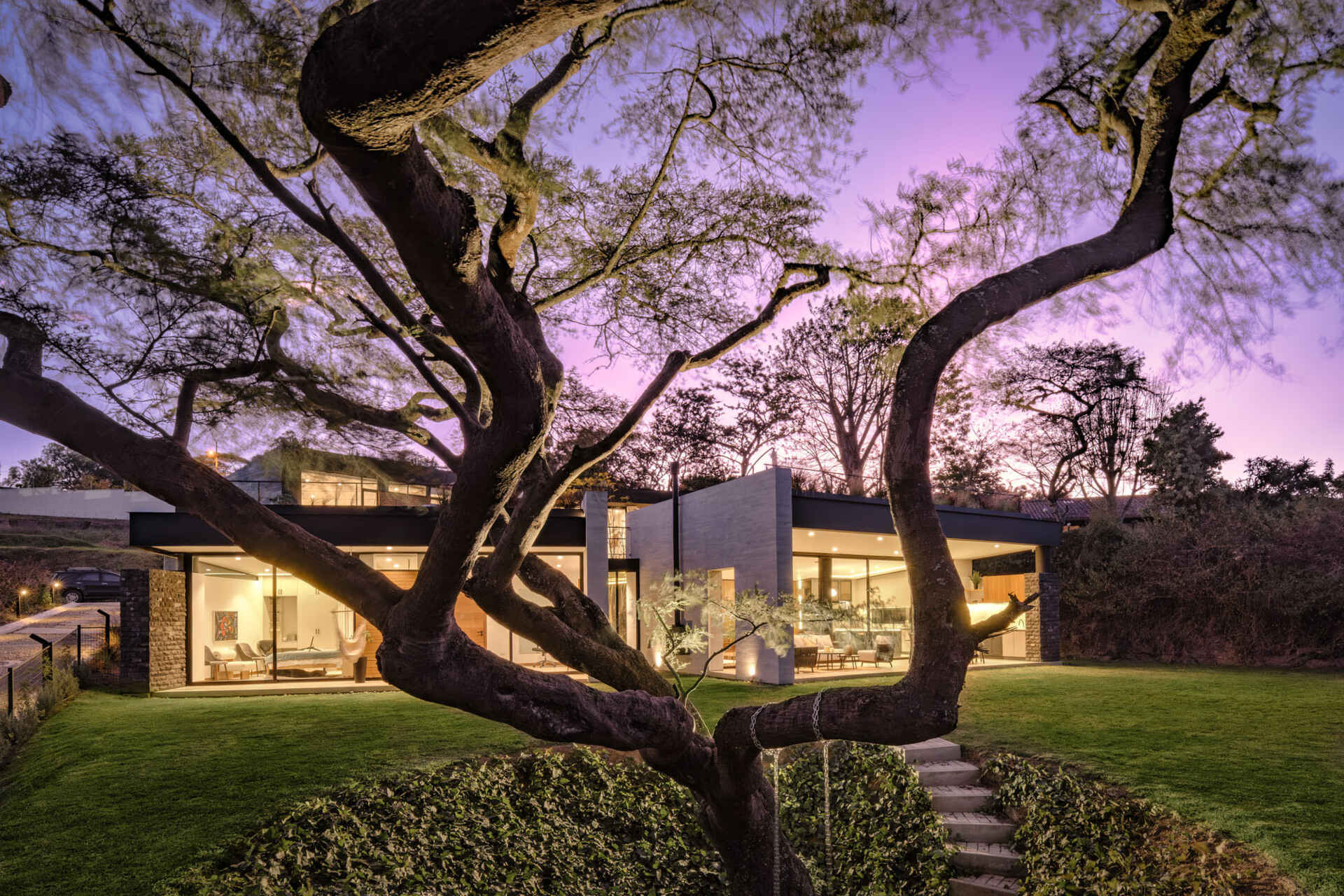 Full-height windows frame garden and treetop views at this contemporary home in Puembo, Ecuador. Slim black frames, wood-clad panels, and projecting roof edges create a facade that reveals the interiors as evening falls.