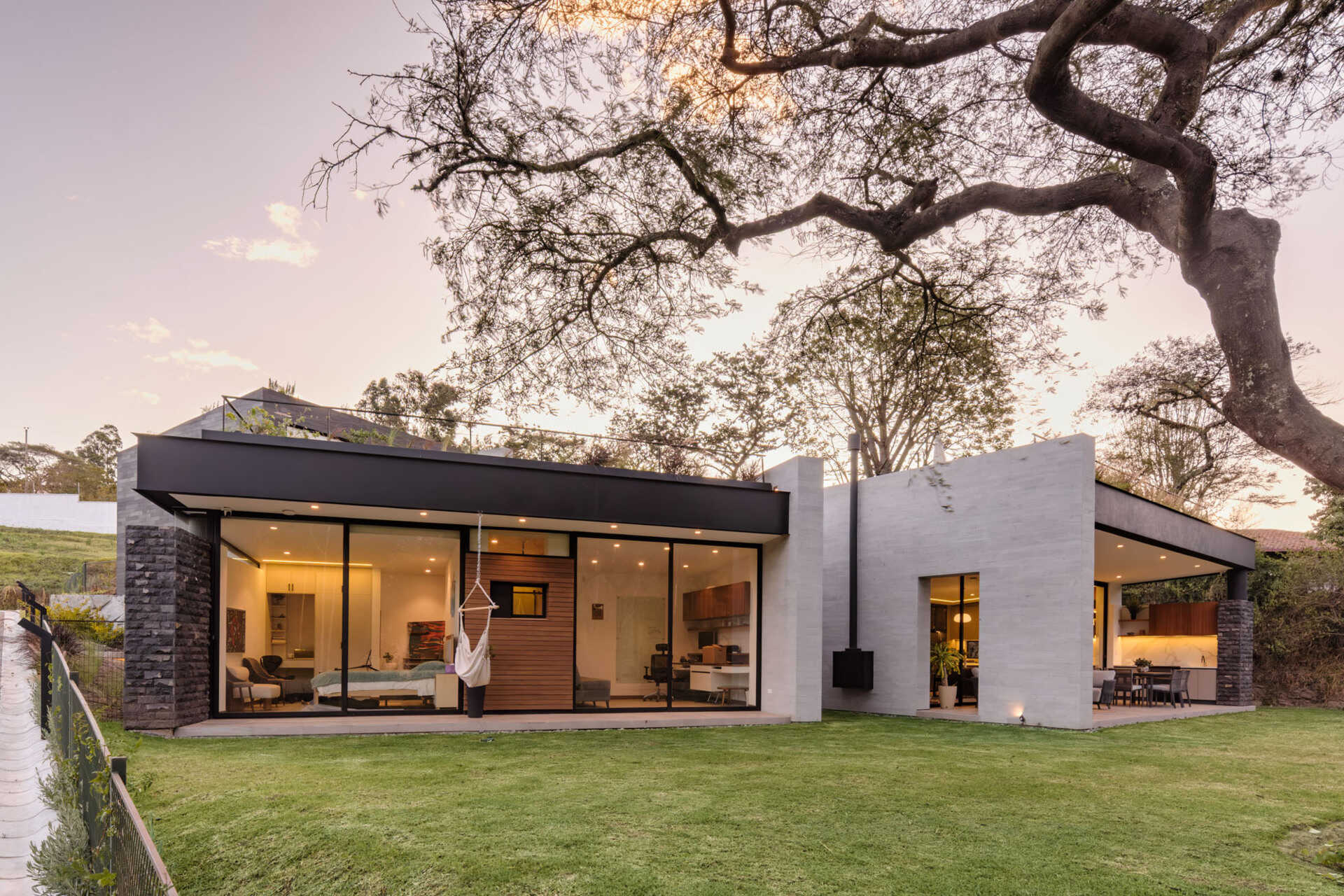Full-height windows frame garden and treetop views at this contemporary home in Puembo, Ecuador. Slim black frames, wood-clad panels, and projecting roof edges create a facade that reveals the interiors as evening falls.