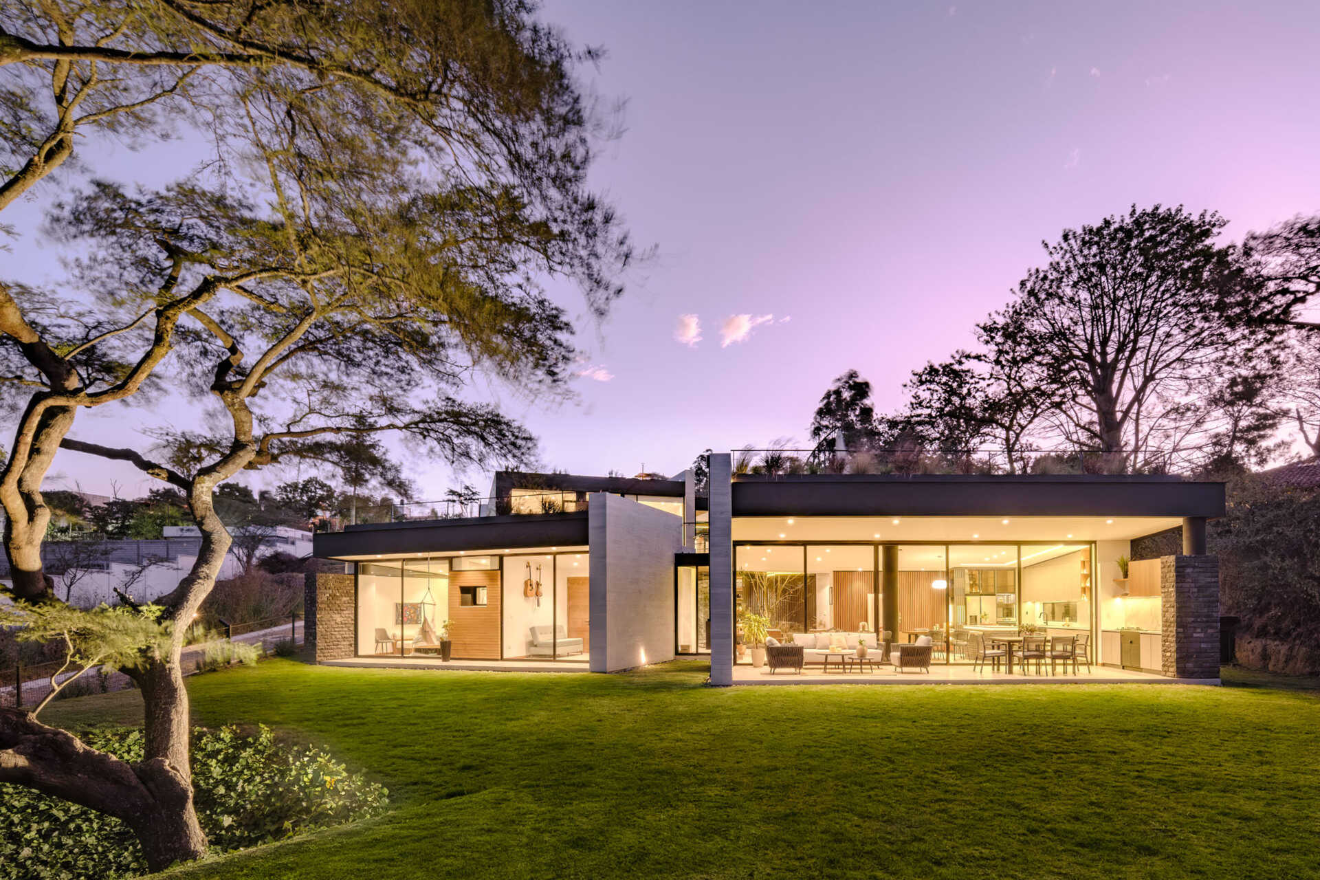 Full-height windows frame garden and treetop views at this contemporary home in Puembo, Ecuador. Slim black frames, wood-clad panels, and projecting roof edges create a facade that reveals the interiors as evening falls.