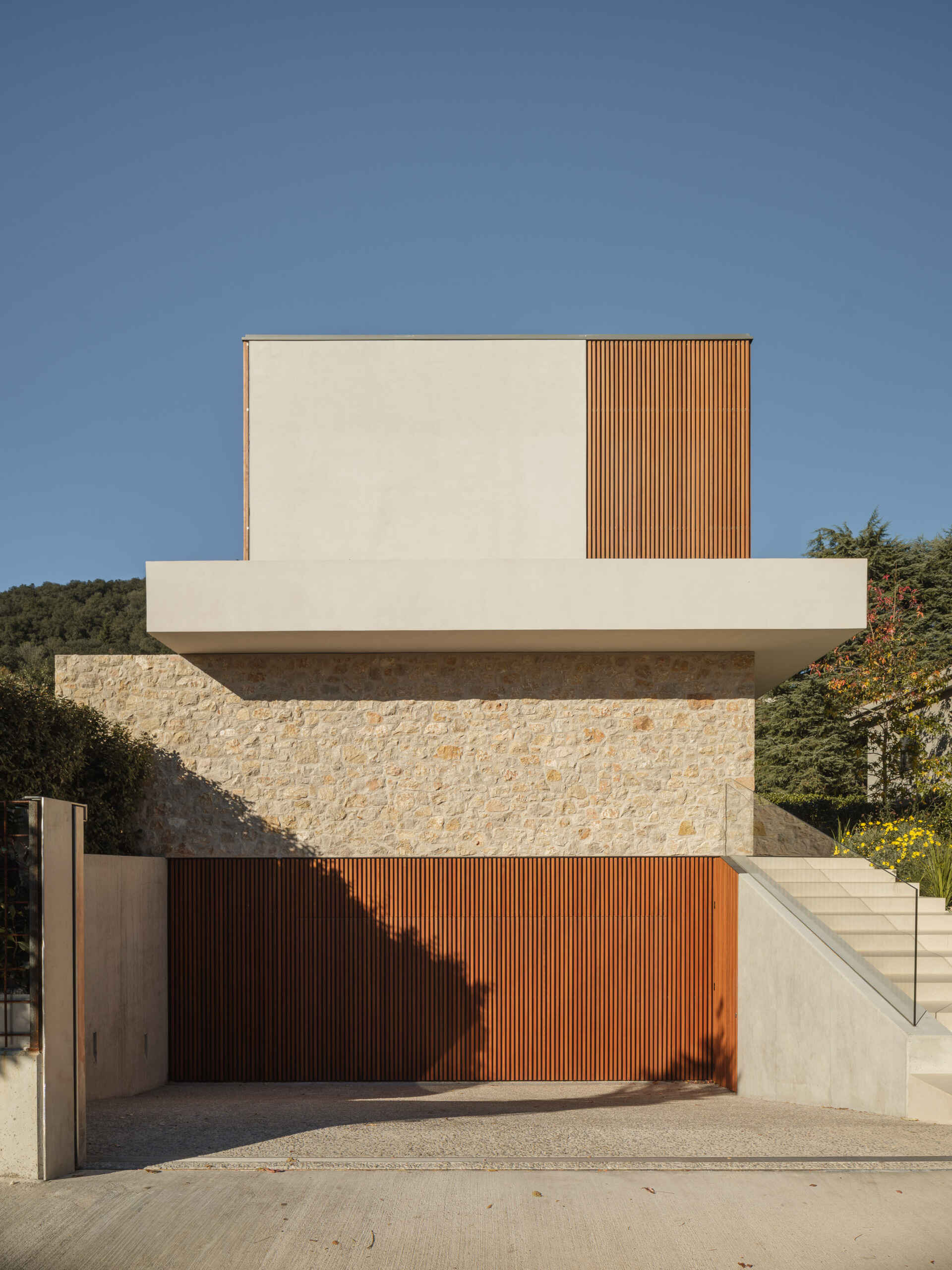 Stone walls, wooden screens, and a projecting concrete slab define this contemporary home in Olot, Spain. Explore House 2194 by Nordest Architecture SLP, with an L-shaped layout, a sheltered garden, and a layered façade.