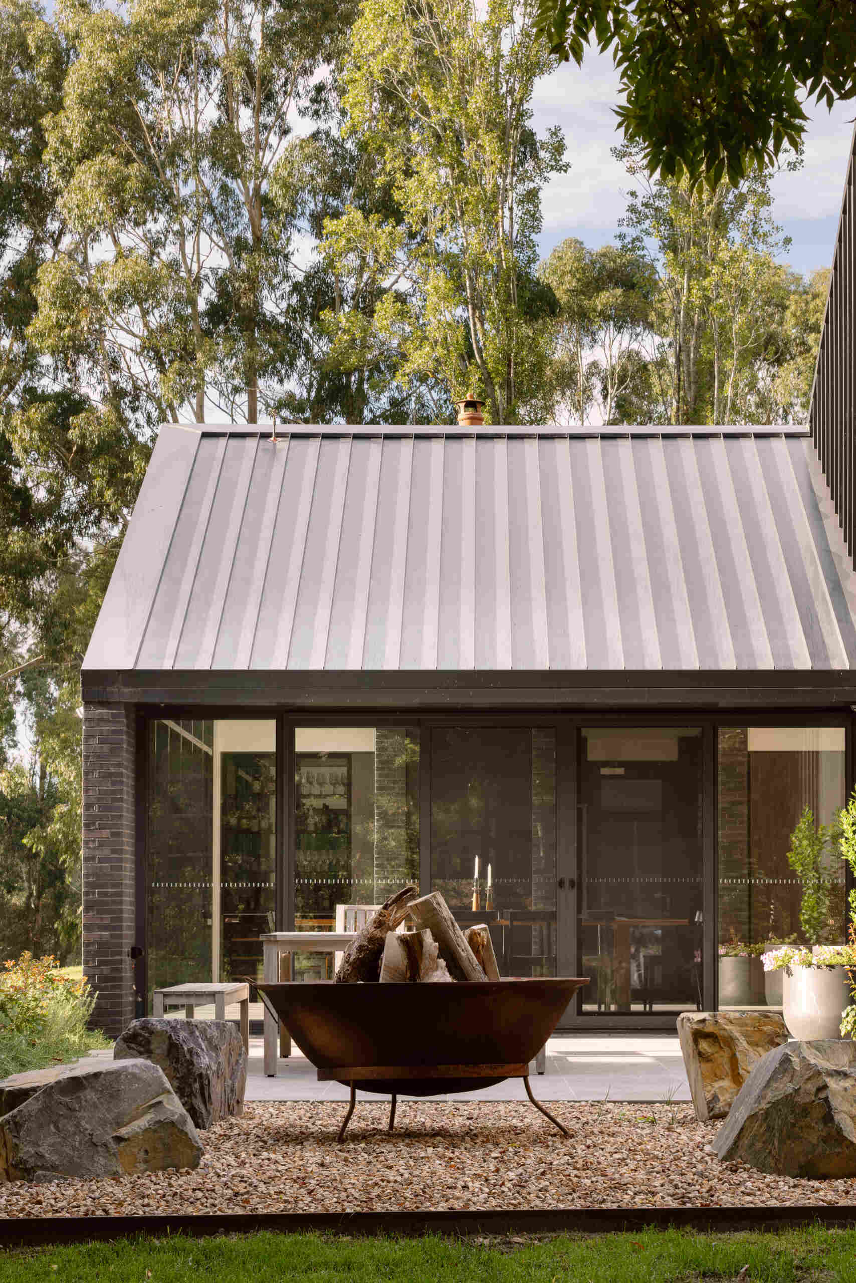 The home's relationship with the landscape continues through its generous areas of glazing and open spaces. A patio furnished with a dining table, and a nearby firepit area, create a casual place to relax outdoors.