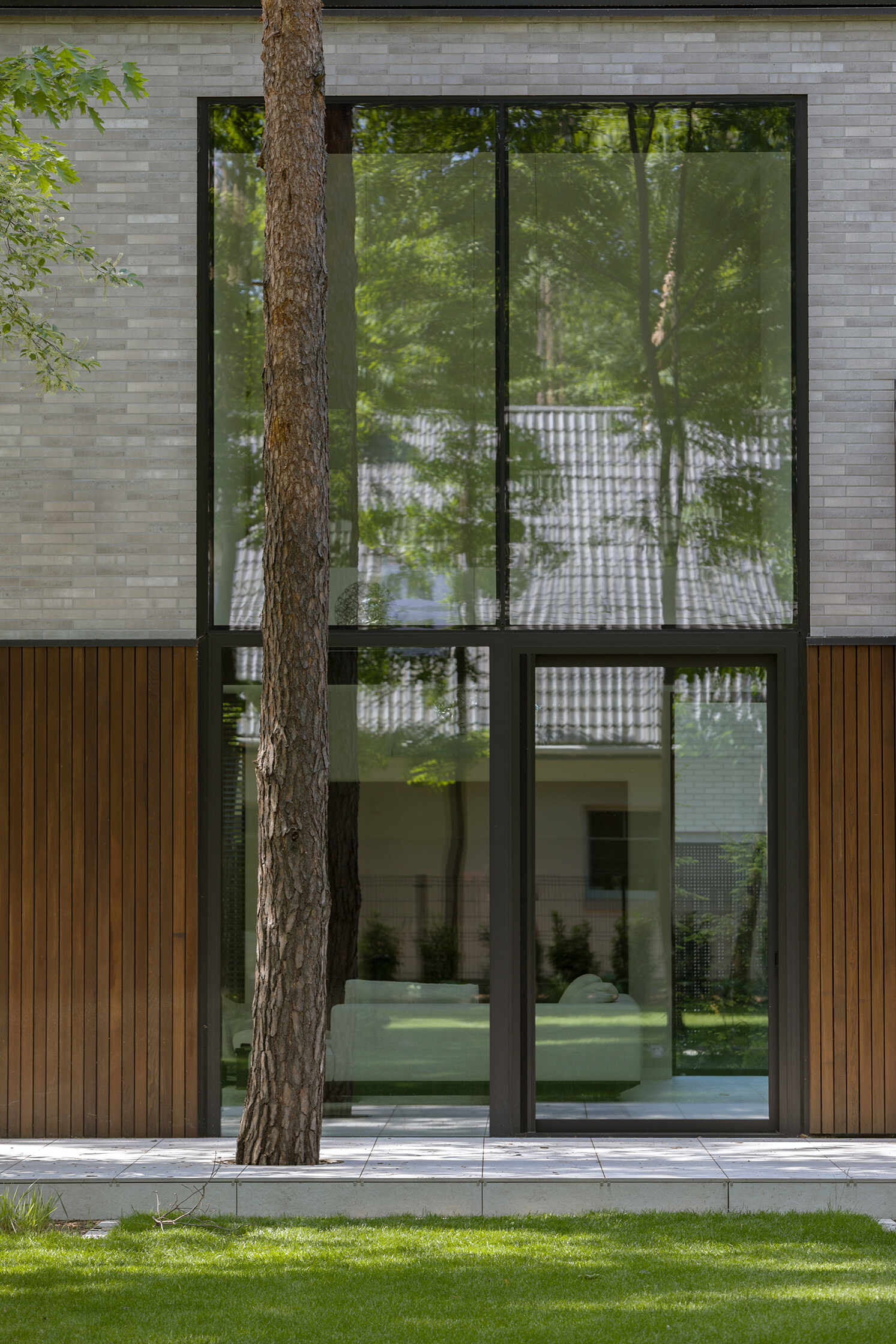 Expansive horizontal glazing and tall vertical windows bring daylight and views of the surrounding pine trees into The Intersecting House near Warsaw, with large living room glazing opening directly onto the terrace.