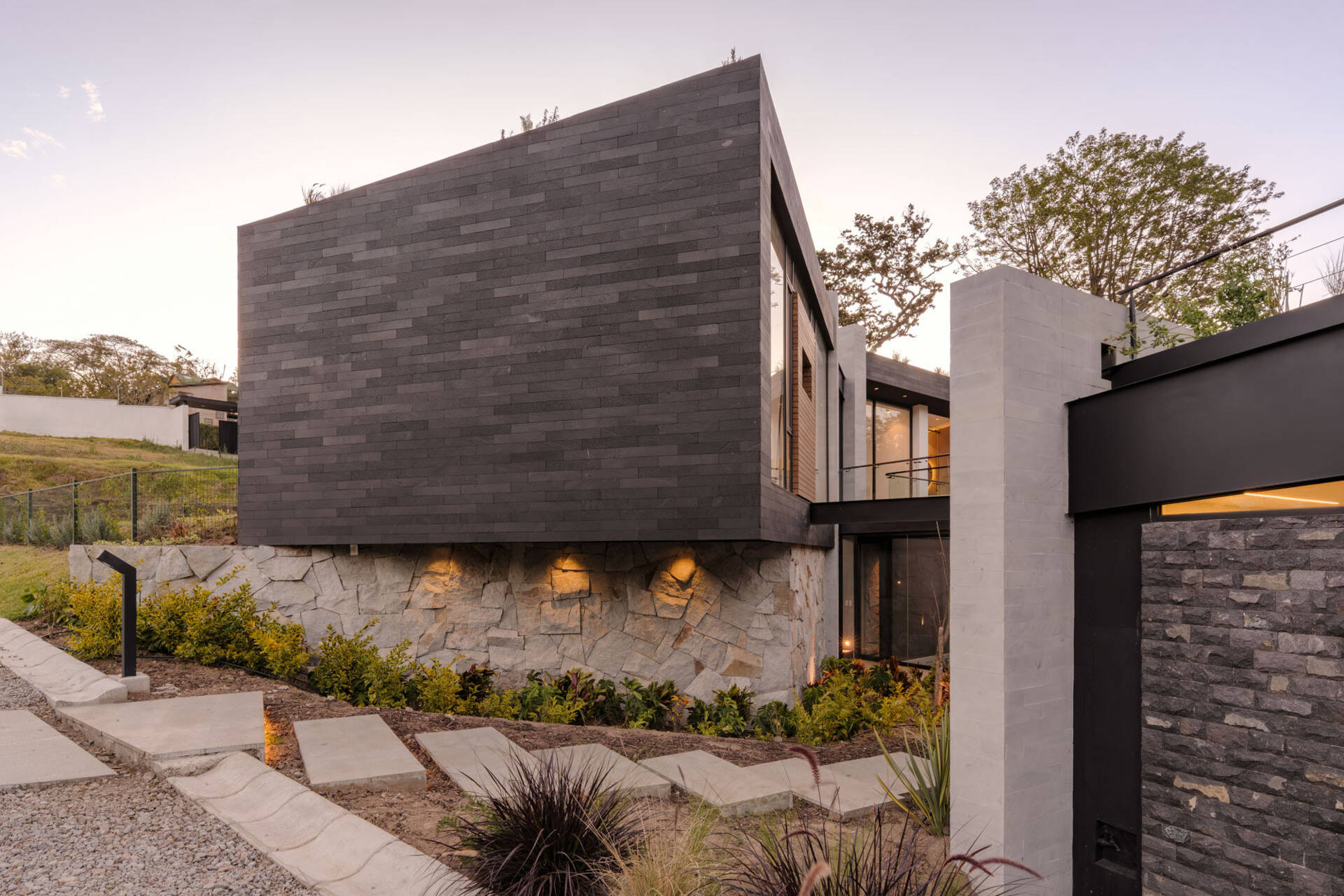 A landscaped entrance in Puembo, Ecuador, pairs staggered concrete stepping slabs with low shrubs, ornamental grasses, and textured stone walls. The path leads to a recessed timber front door with a long black handle and adjacent glazing.
