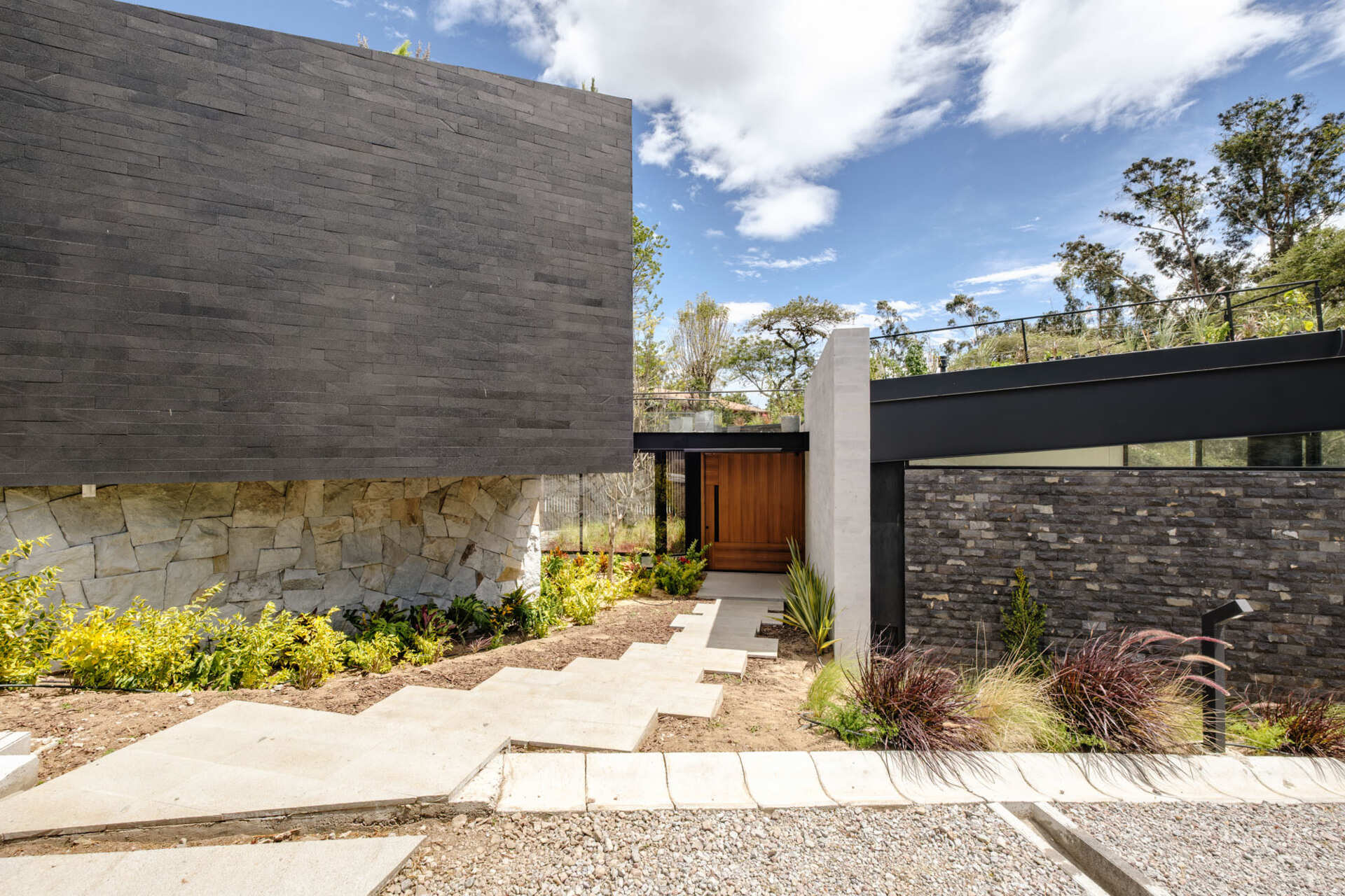 A landscaped entrance in Puembo, Ecuador, pairs staggered concrete stepping slabs with low shrubs, ornamental grasses, and textured stone walls. The path leads to a recessed timber front door with a long black handle and adjacent glazing.