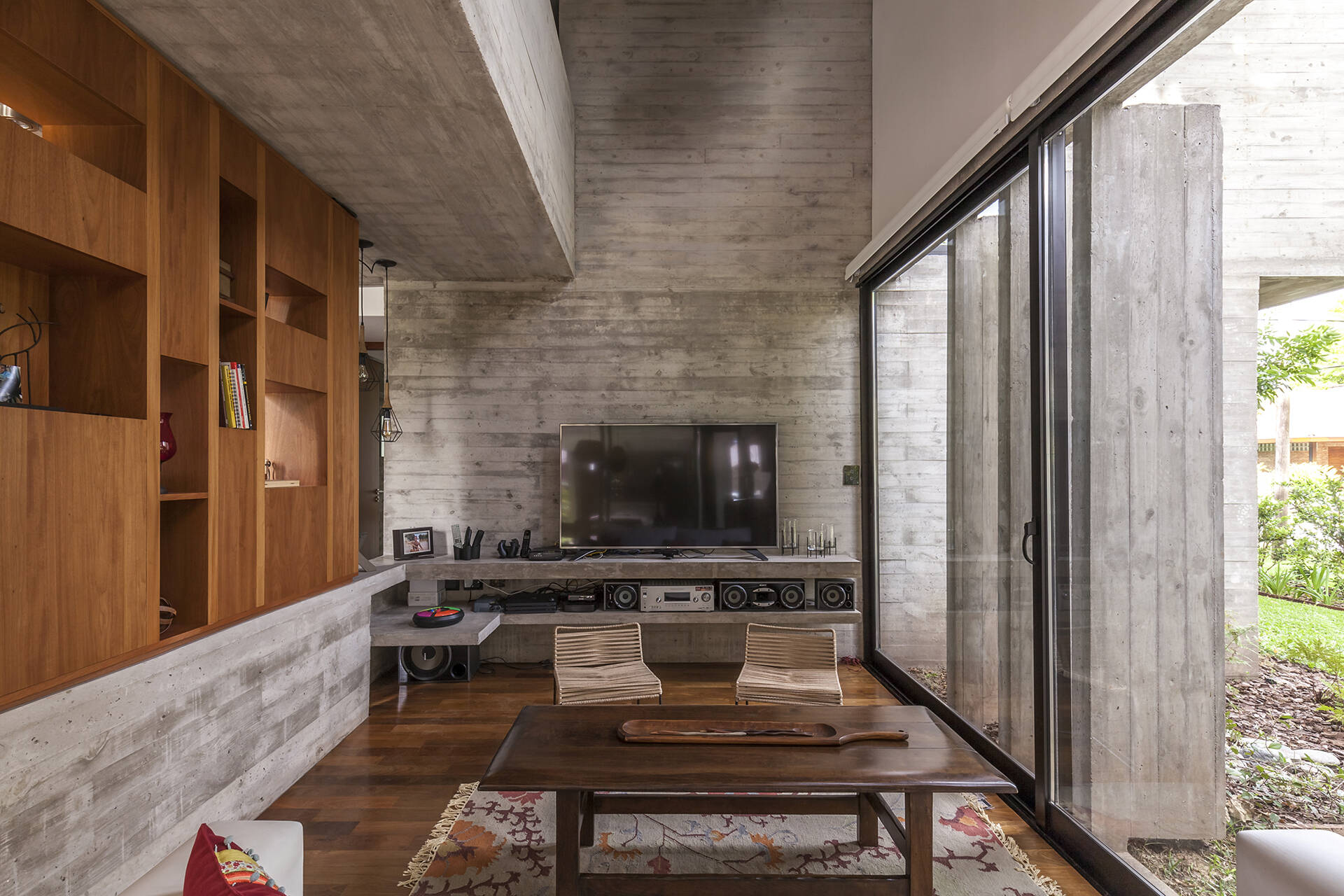 Explore the modern living room of Casa Aranzazu, where exposed concrete, warm wood shelving, large windows and a built-in fireplace create a clean yet inviting interior.