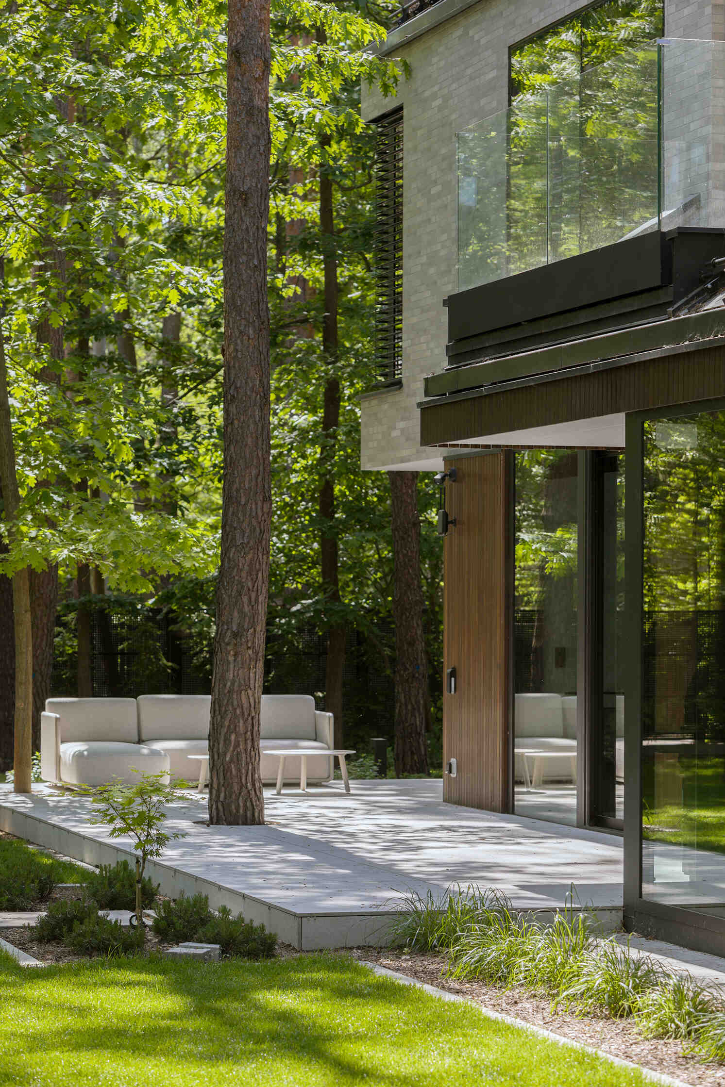 The covered west-facing terrace at The Intersecting House creates a sheltered outdoor space beneath the upper volume. Large openings connect the terrace with the double-height living room and garden.