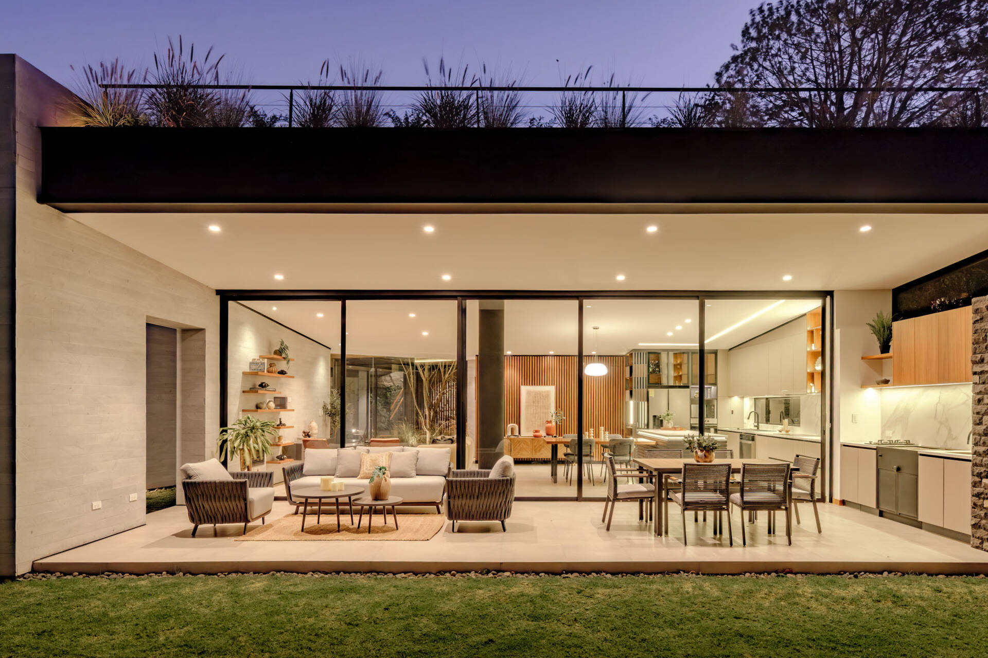 A covered patio opens directly from the living and dining rooms through wide sliding glass doors. Woven outdoor seating, a dining table, and a built-in grill and sink create a sheltered space overlooking the lawn at this contemporary home in Ecuador.