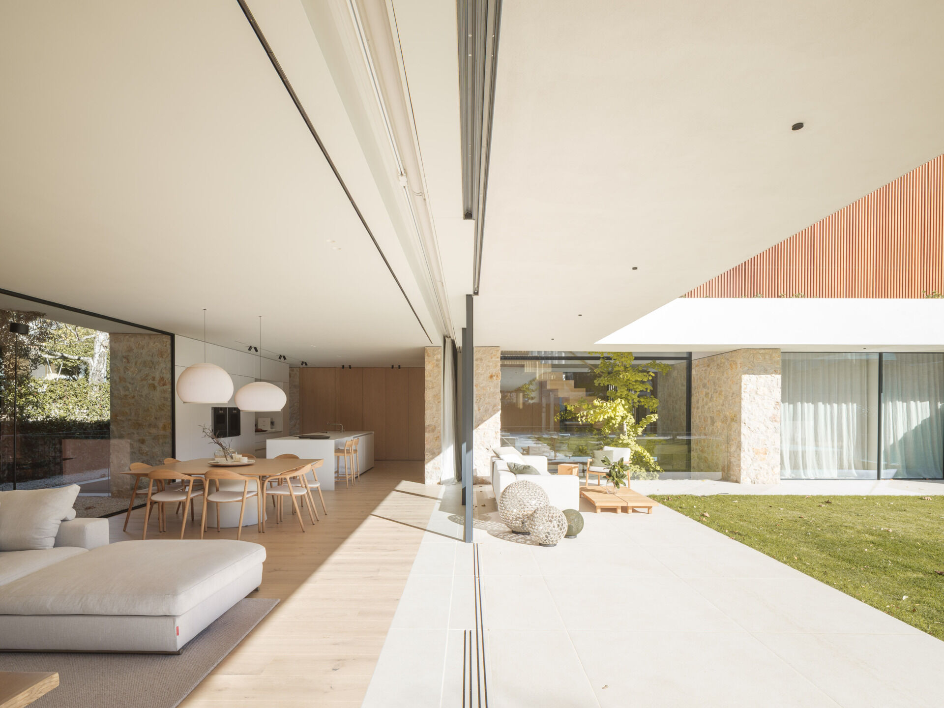 A shaded patio, timber outdoor furniture, central lawn, and pool create an inviting backyard at House 2194 in Olot, Spain. Sliding glass doors open the living and dining spaces directly onto the garden.