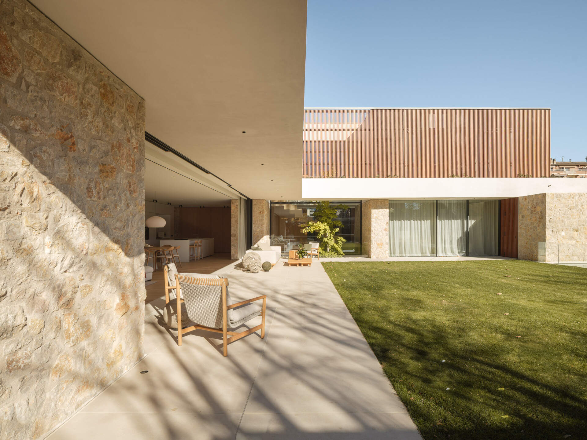 A shaded patio, timber outdoor furniture, central lawn, and pool create an inviting backyard at House 2194 in Olot, Spain. Sliding glass doors open the living and dining spaces directly onto the garden.