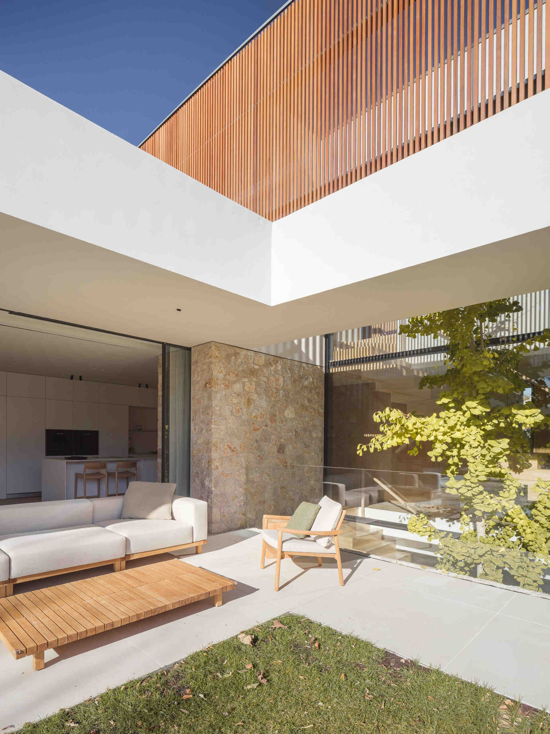 A shaded patio, timber outdoor furniture, central lawn, and pool create an inviting backyard at House 2194 in Olot, Spain. Sliding glass doors open the living and dining spaces directly onto the garden.