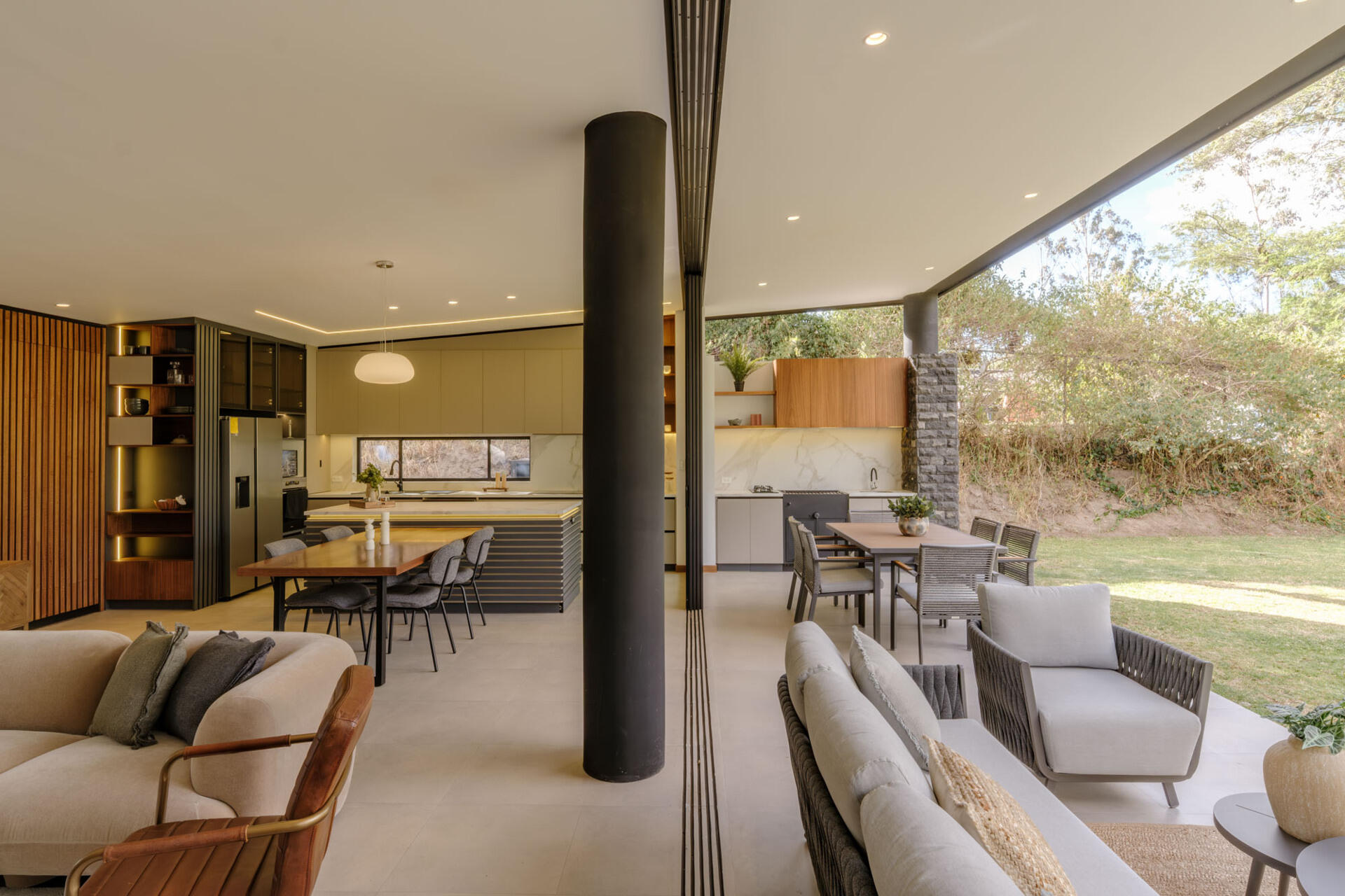 A covered patio opens directly from the living and dining rooms through wide sliding glass doors. Woven outdoor seating, a dining table, and a built-in grill and sink create a sheltered space overlooking the lawn at this contemporary home in Ecuador.