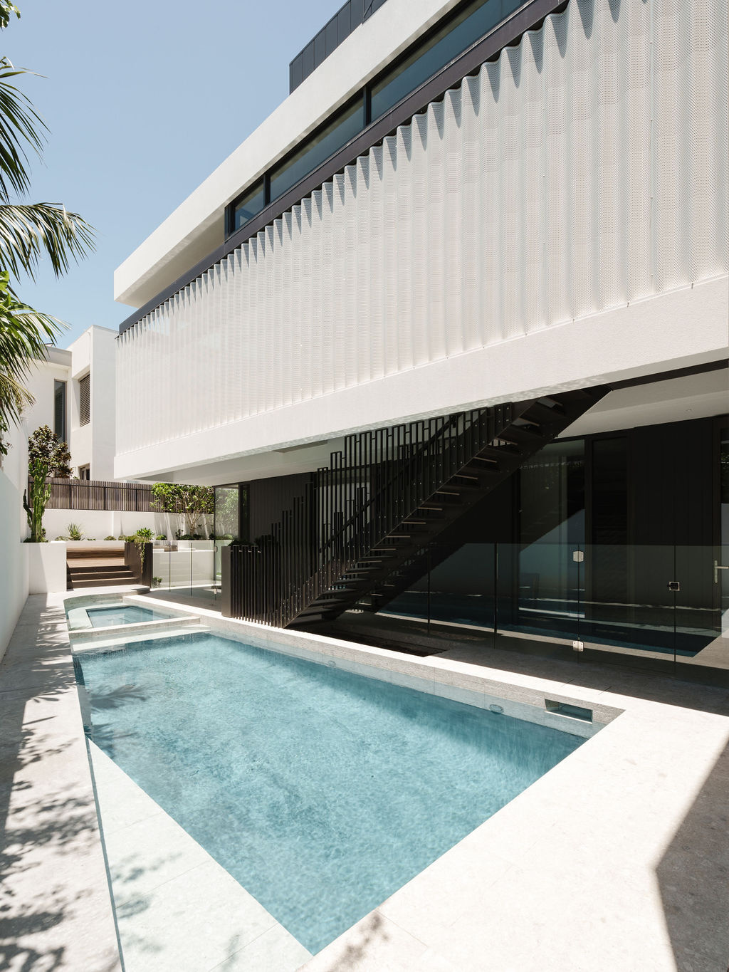 The pool at Mezzanine House sits alongside the architecture, with pale paving, glass walls, a black steel staircase and a textured perforated facade.