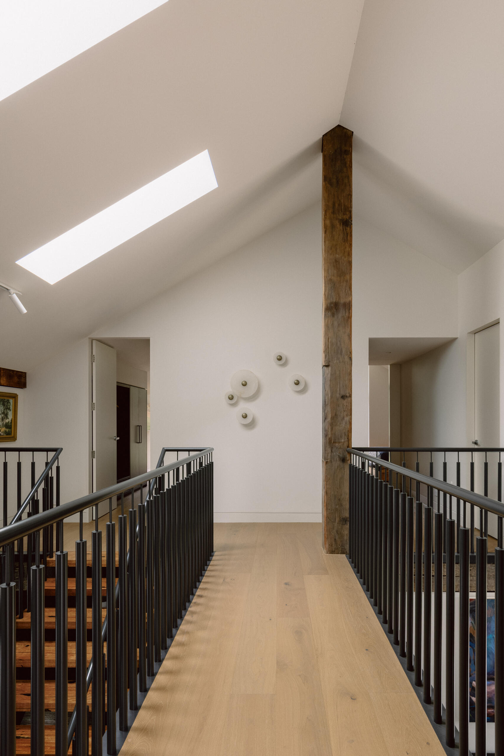 A light-filled upper bridge walkway in Yarra Valley House featuring pale timber flooring, black metal railings, an exposed timber post and a dramatic skylight.