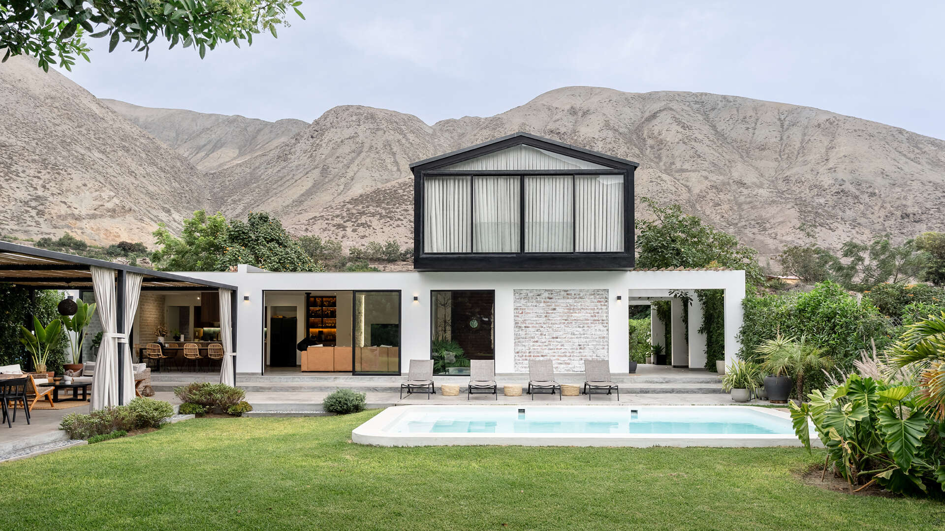Tall black-framed windows line the white ground floor of Casa Berna in Peru, with ornamental grasses below and a dark upper volume rising behind them.