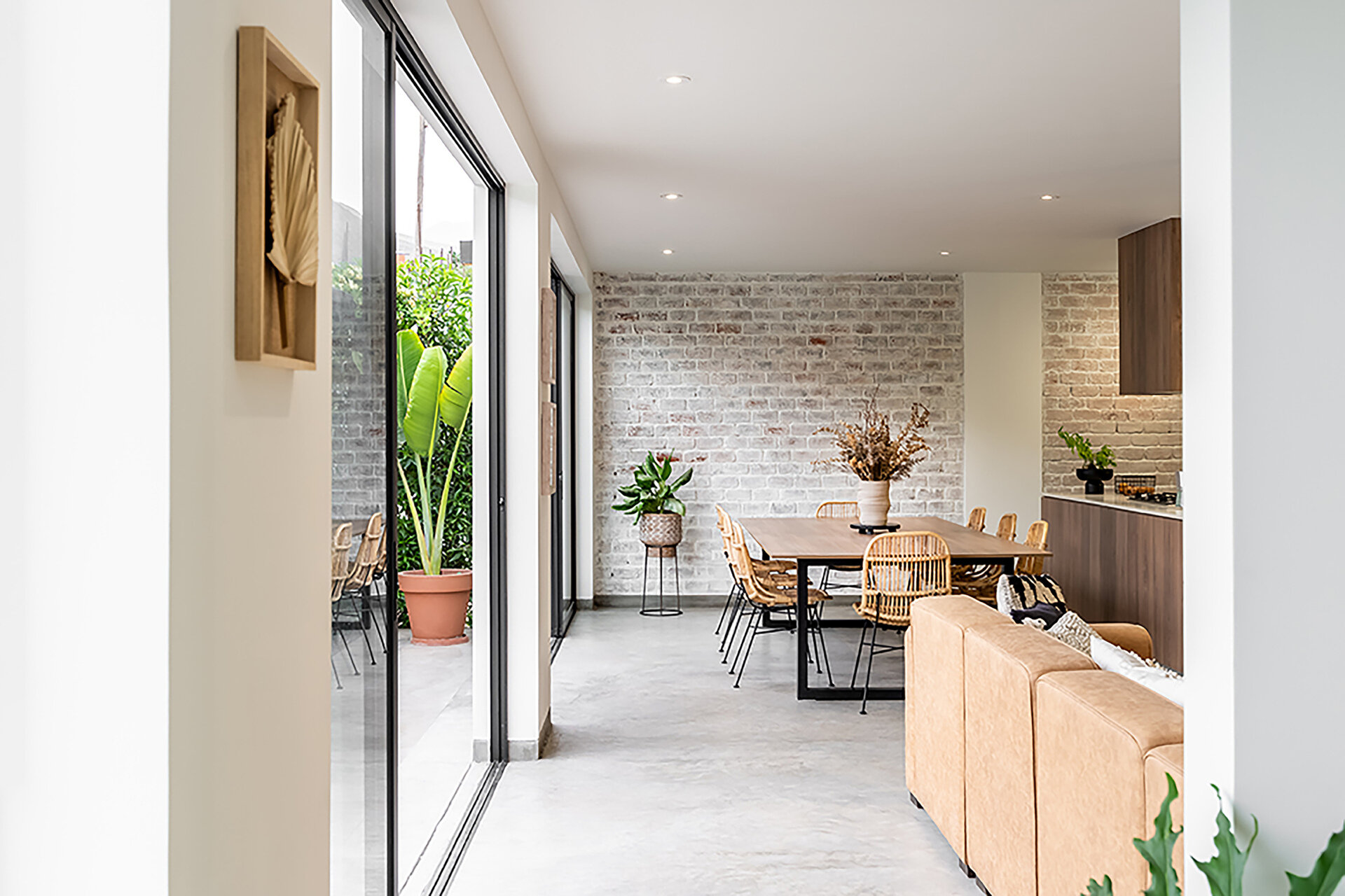 At Casa Berna, a timber dining table and woven chairs sit beside garden-facing glass doors, with a lime-washed brick wall behind them.