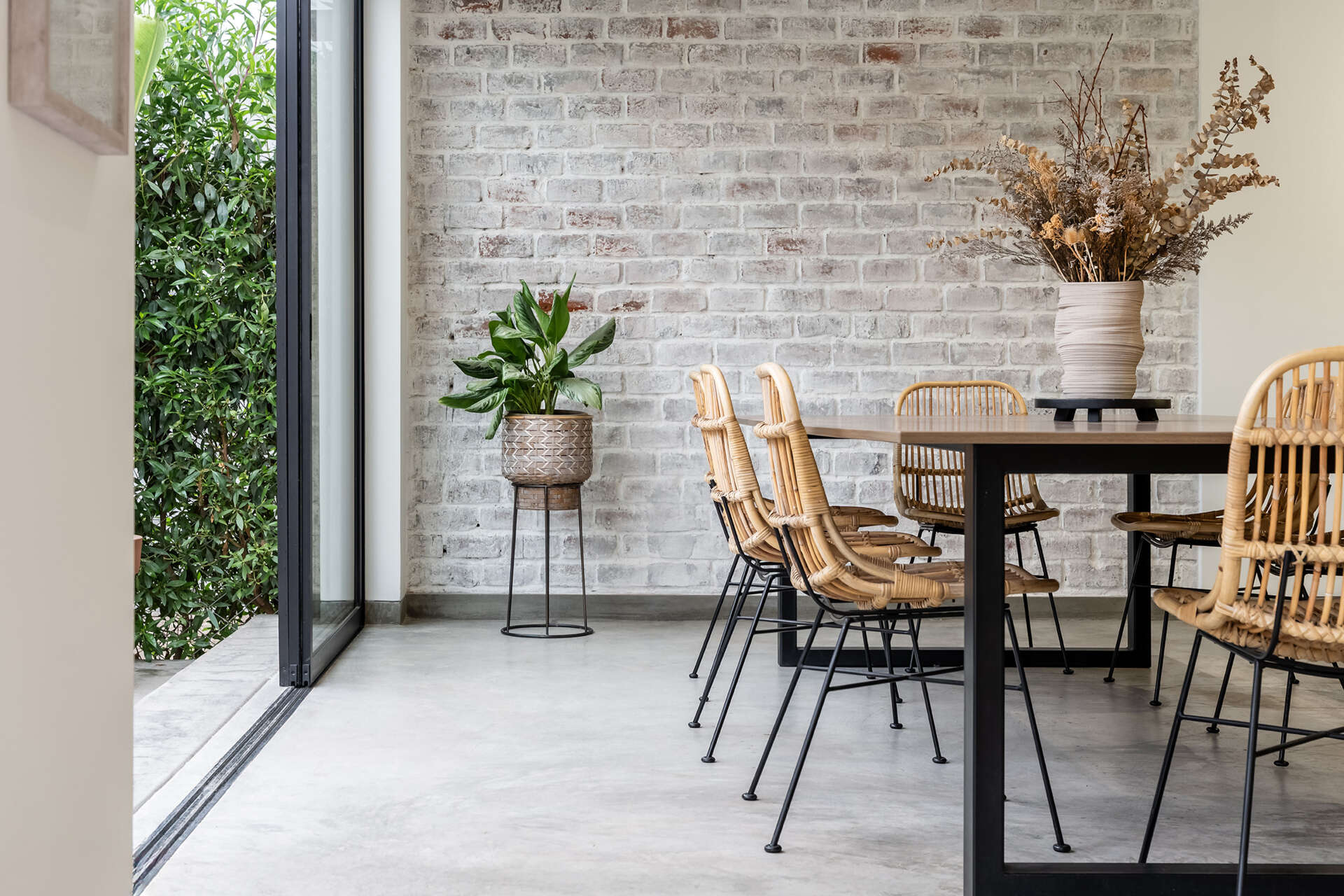 At Casa Berna, a timber dining table and woven chairs sit beside garden-facing glass doors, with a lime-washed brick wall behind them.