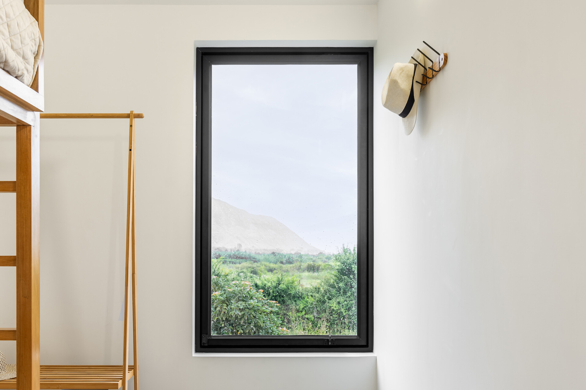 A tall window frames greenery and distant hills in Casa Berna’s kids’ bedroom, alongside a timber bunk bed and simple clothes rail.
