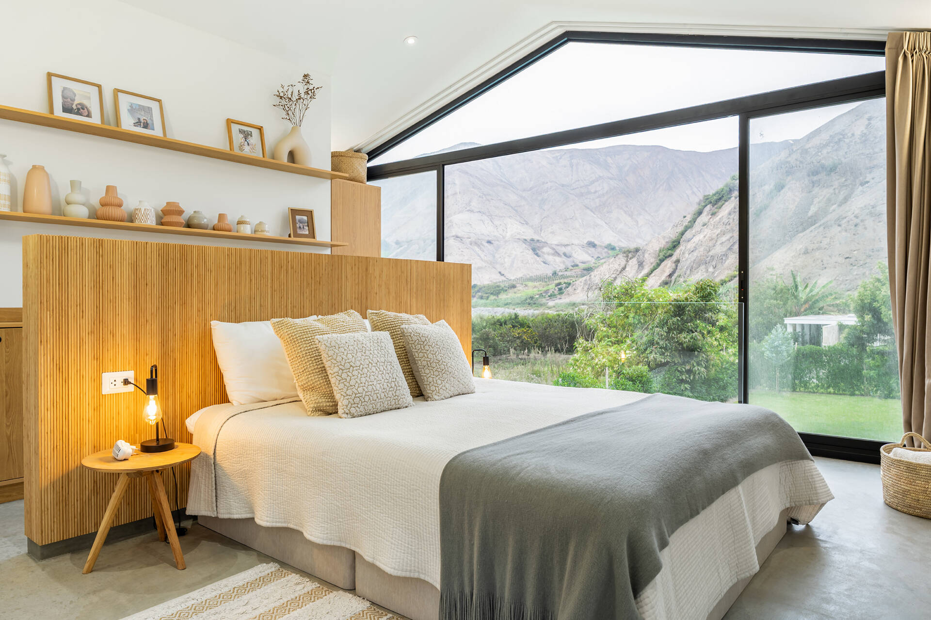 Casa Berna’s primary bedroom has a roofline-shaped window overlooking Peru’s valley hills, alongside a slatted timber headboard, open shelves and soft neutral bedding.