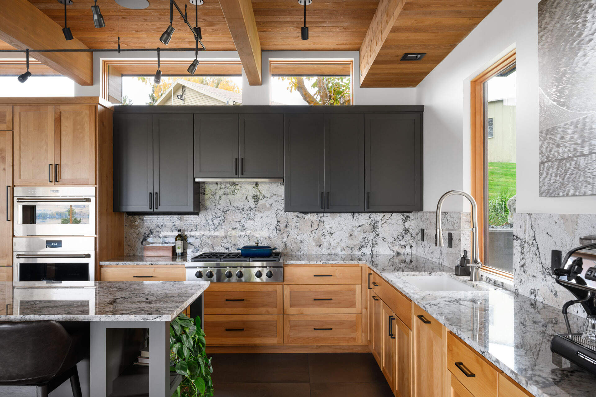 Natural wood cabinetry, dark grey storage, veined stone surfaces, and exposed timber beams create a warm modern kitchen with plenty of visual texture.