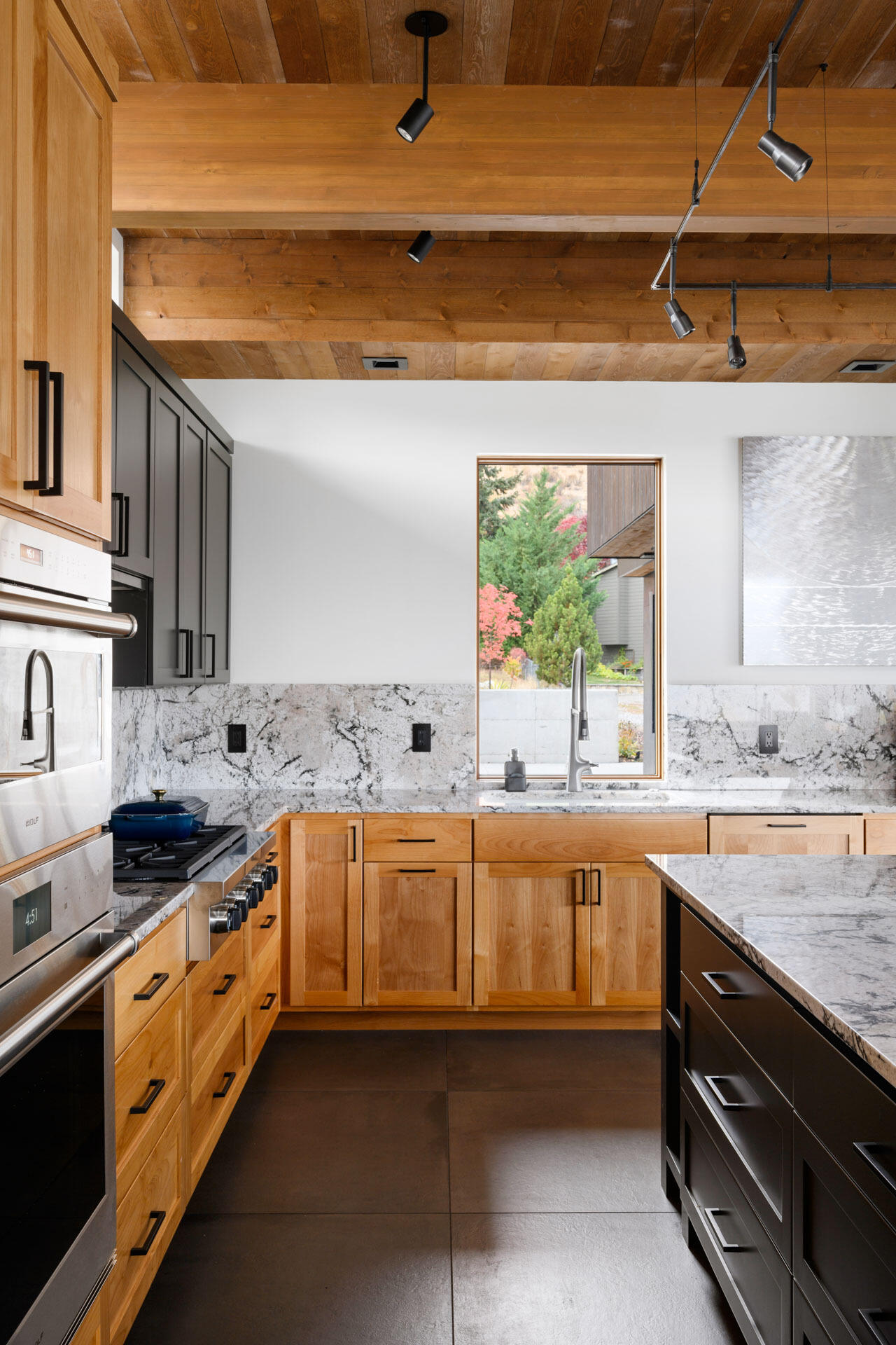 Natural wood cabinetry, dark grey storage, veined stone surfaces, and exposed timber beams create a warm modern kitchen with plenty of visual texture.