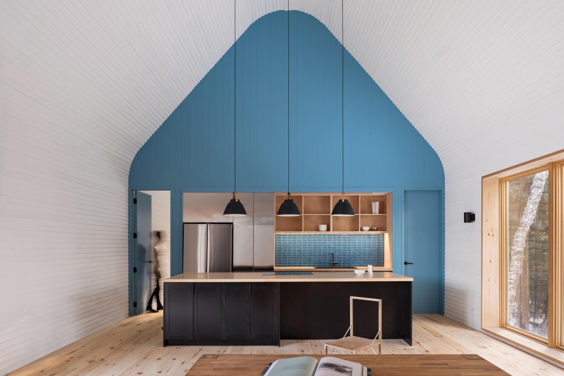 A modern cottage kitchen featuring a blue painted gable wall, matching tile backsplash, timber shelving, a dark kitchen island, and large windows overlooking the forest.