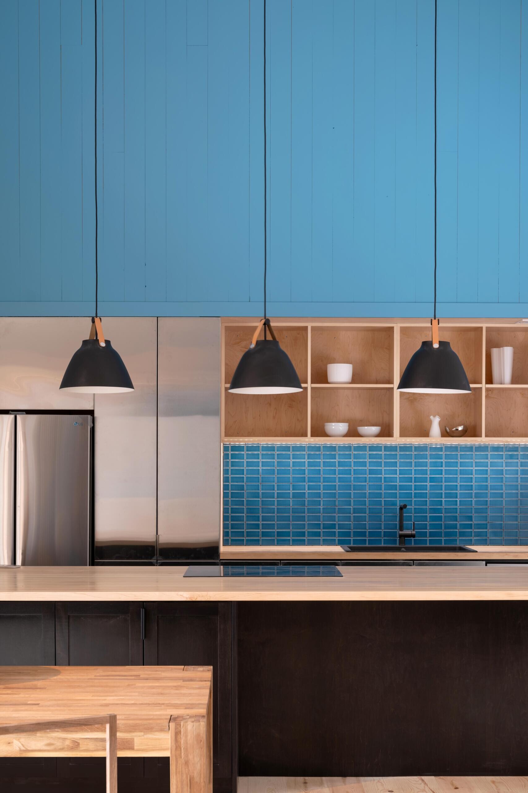 A modern cottage kitchen featuring a blue painted gable wall, matching tile backsplash, timber shelving, a dark kitchen island, and large windows overlooking the forest.