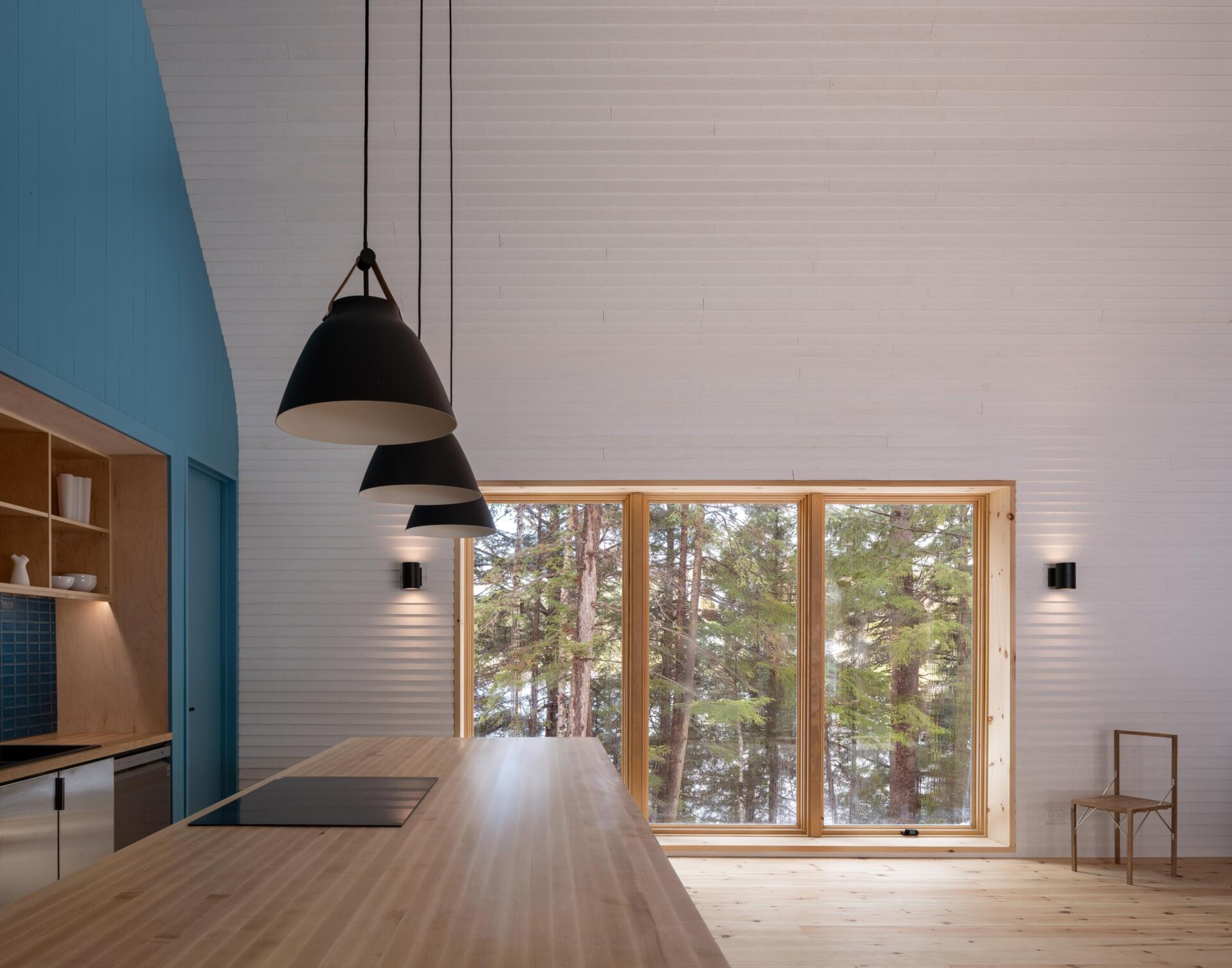 A modern cottage kitchen featuring a blue painted gable wall, matching tile backsplash, timber shelving, a dark kitchen island, and large windows overlooking the forest.