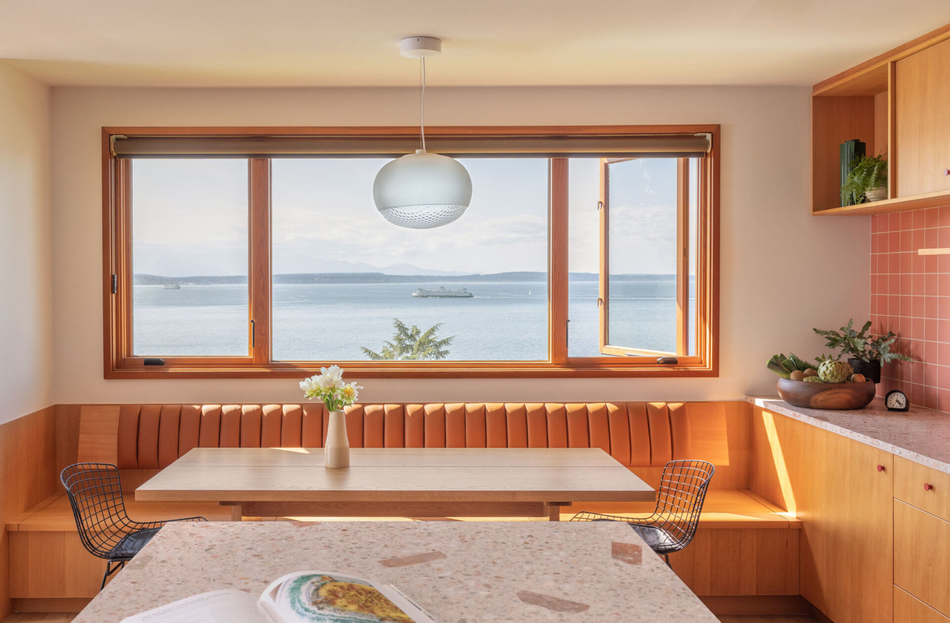 Tour the remodeled kitchen and dining area, where SHED flipped the original layout so a built-in banquette now faces Puget Sound, paired with warm timber cabinetry, pink Concrete Collaborative counters and a Zia Tile backsplash.