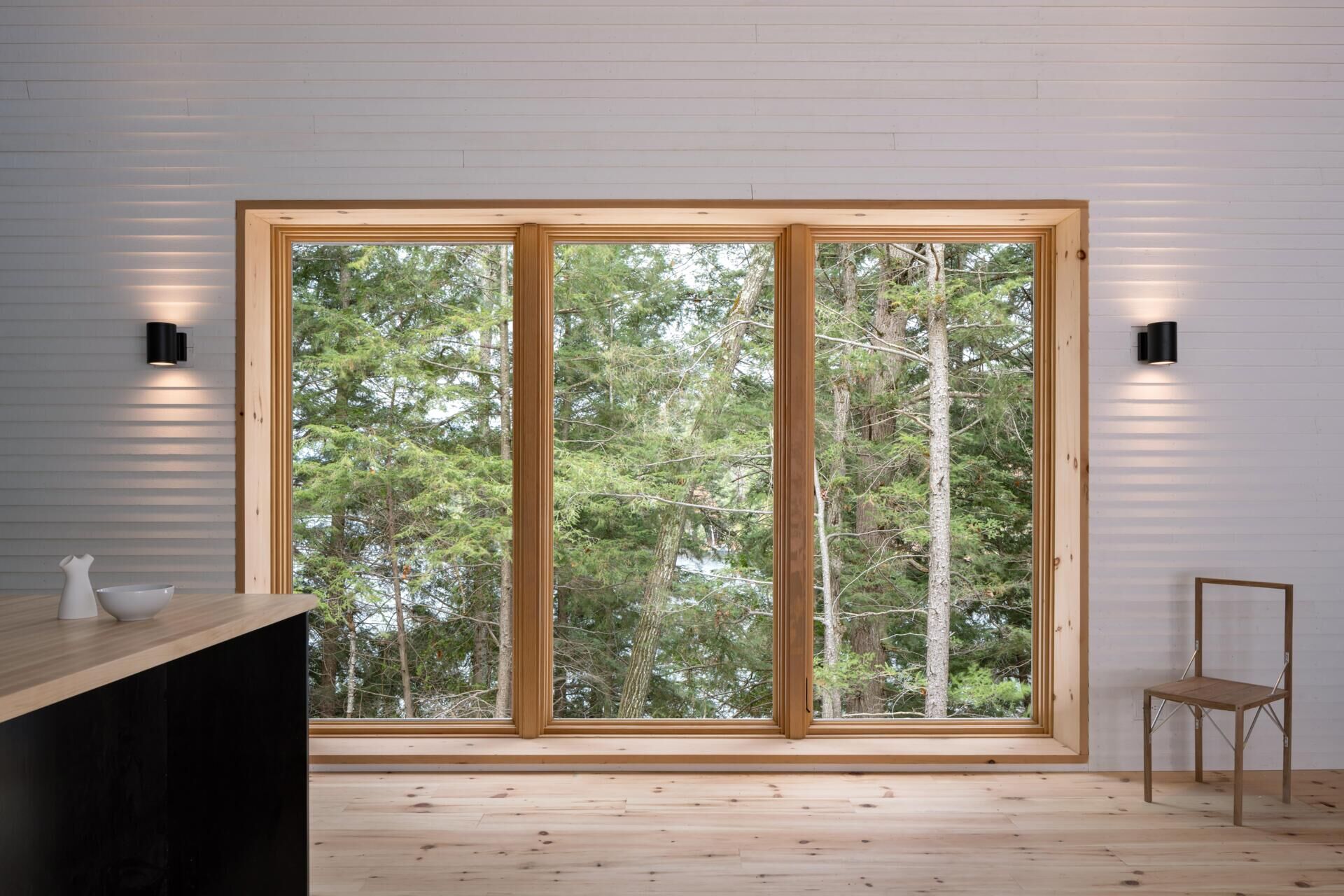 A modern cottage kitchen featuring a blue painted gable wall, matching tile backsplash, timber shelving, a dark kitchen island, and large windows overlooking the forest.