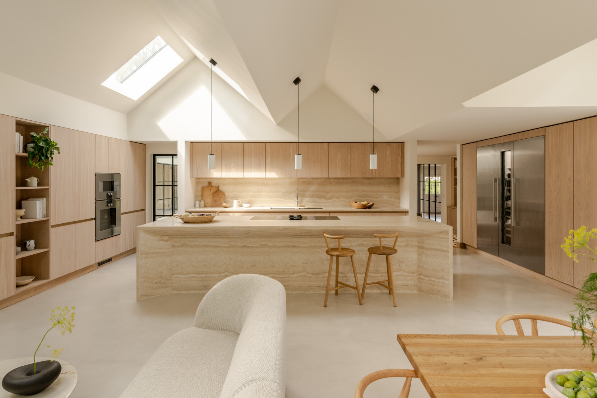 Discover the spacious kitchen at Thirsk Cottage, with pale wood cabinetry, a travertine island, microcement flooring and skylights beneath the dramatic vaulted roof.