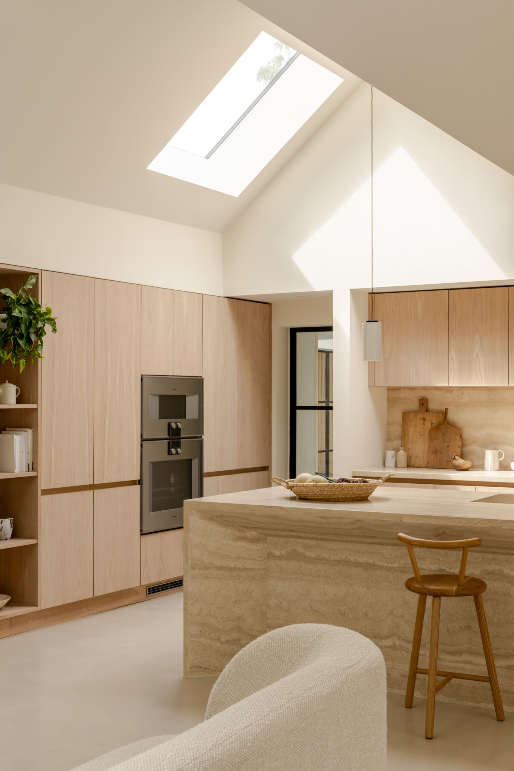 Discover the spacious kitchen at Thirsk Cottage, with pale wood cabinetry, a travertine island, microcement flooring and skylights beneath the dramatic vaulted roof.