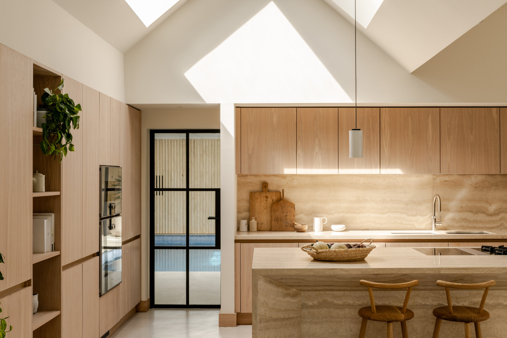 Discover the spacious kitchen at Thirsk Cottage, with pale wood cabinetry, a travertine island, microcement flooring and skylights beneath the dramatic vaulted roof.